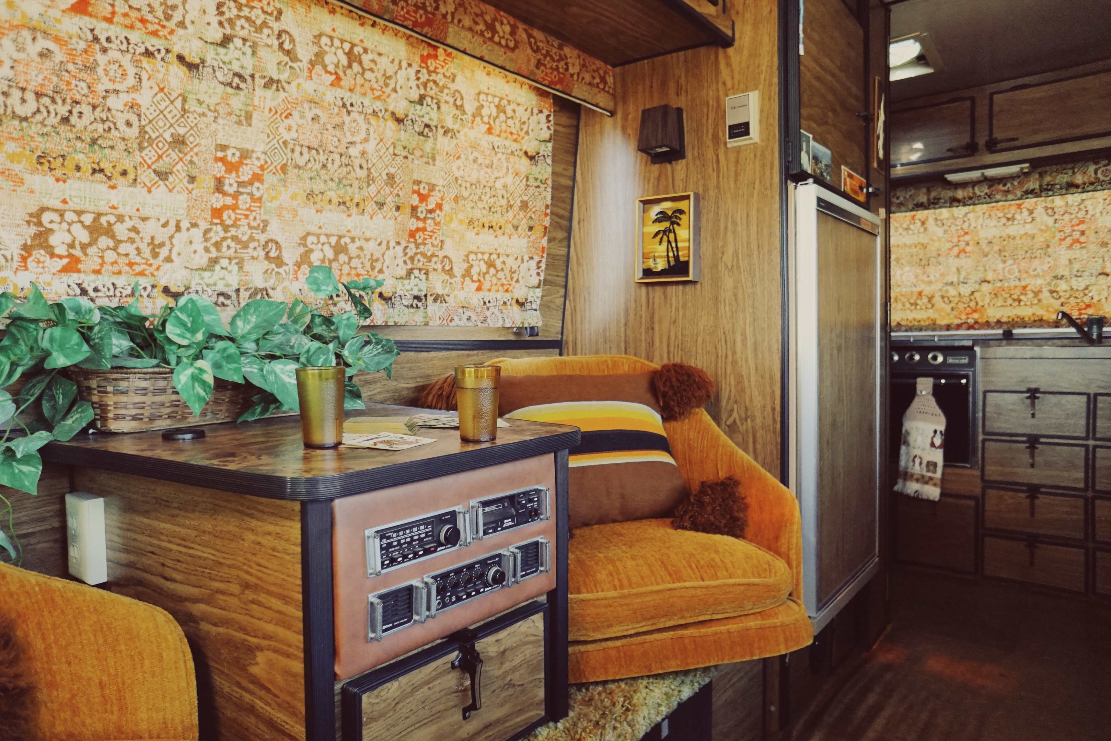 A cozy interior of a vintage camper van featuring an orange armchair, a wooden table with a radio, and patterned curtains.