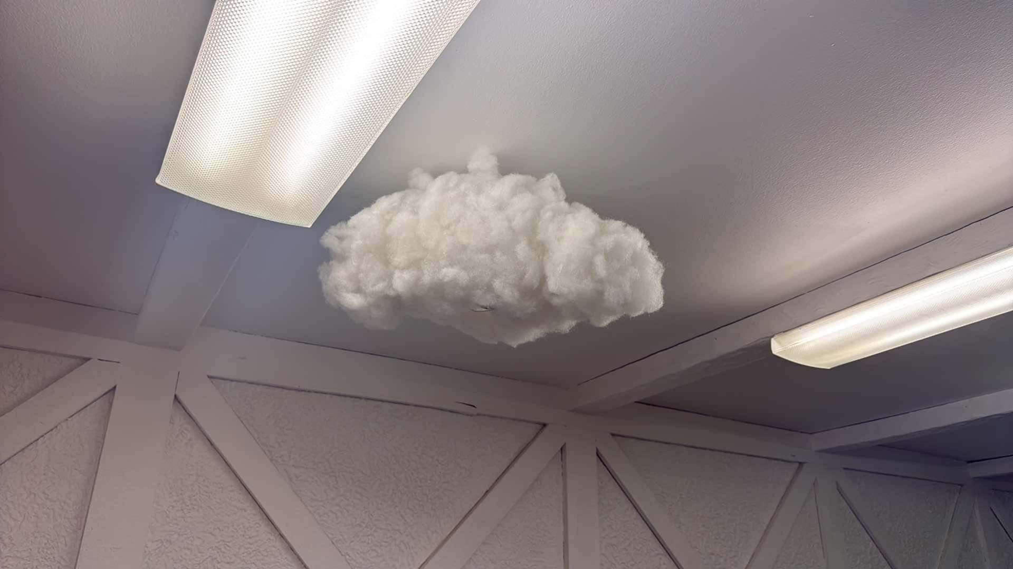 A fluffy, white cloud installation is mounted on the ceiling of a room with a textured wall and fluorescent lights.