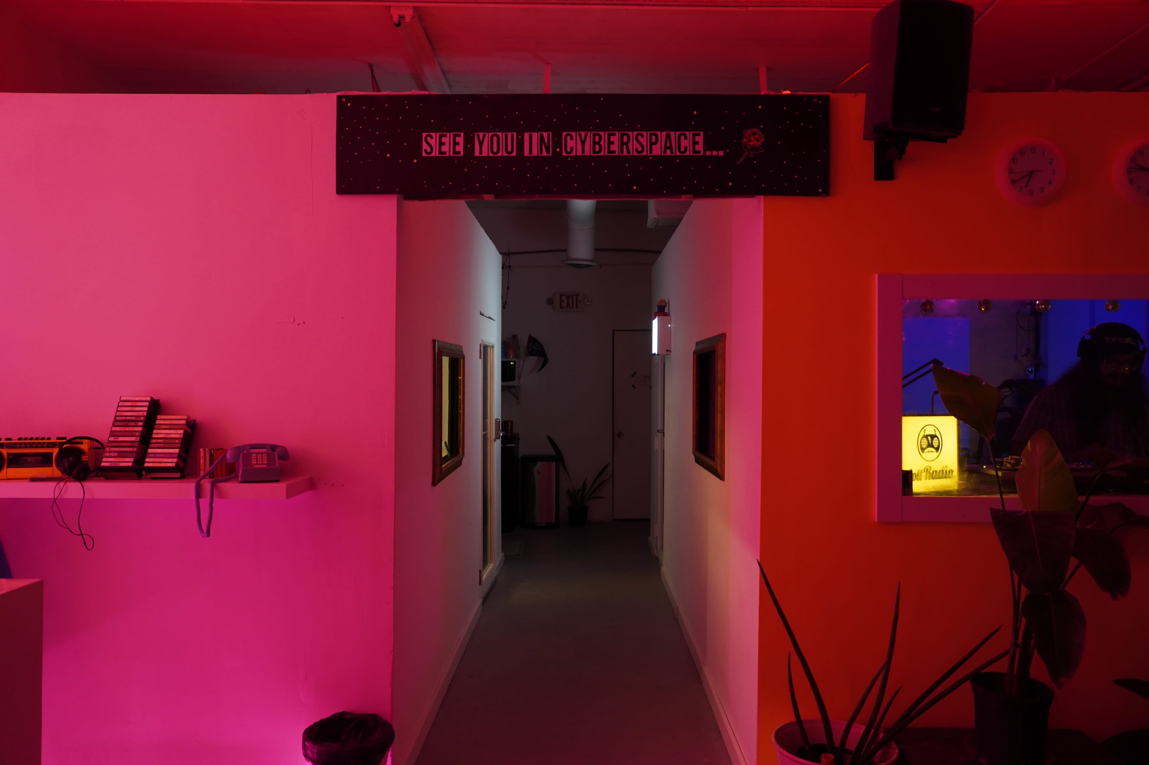 A corridor is illuminated with pink and orange lighting, featuring a sign reading "SEE YOU IN CYBERSPACE" and a small window showing a person working inside.