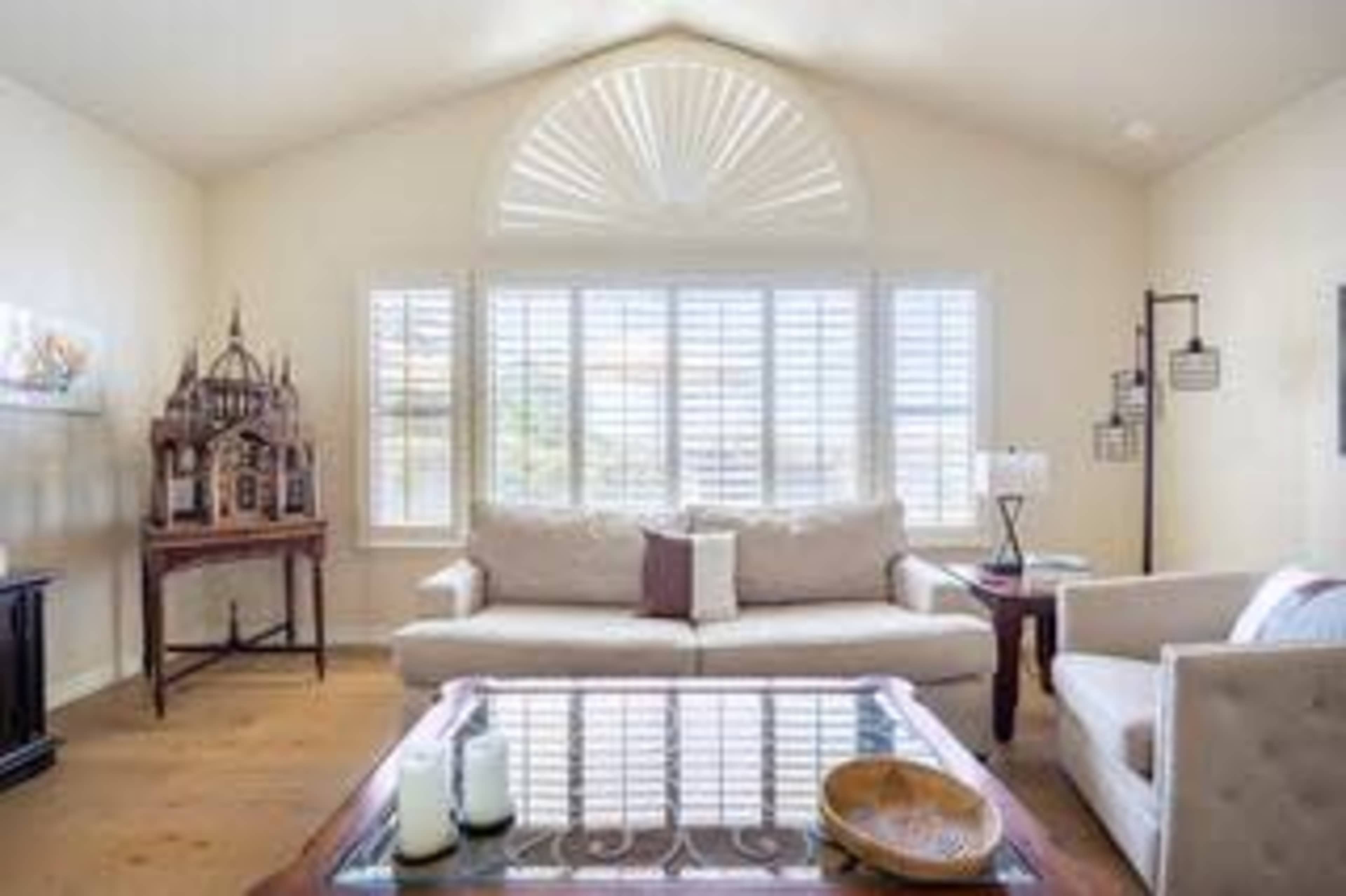A bright living room features a large sofa, a coffee table, and windows adorned with plantation shutters.