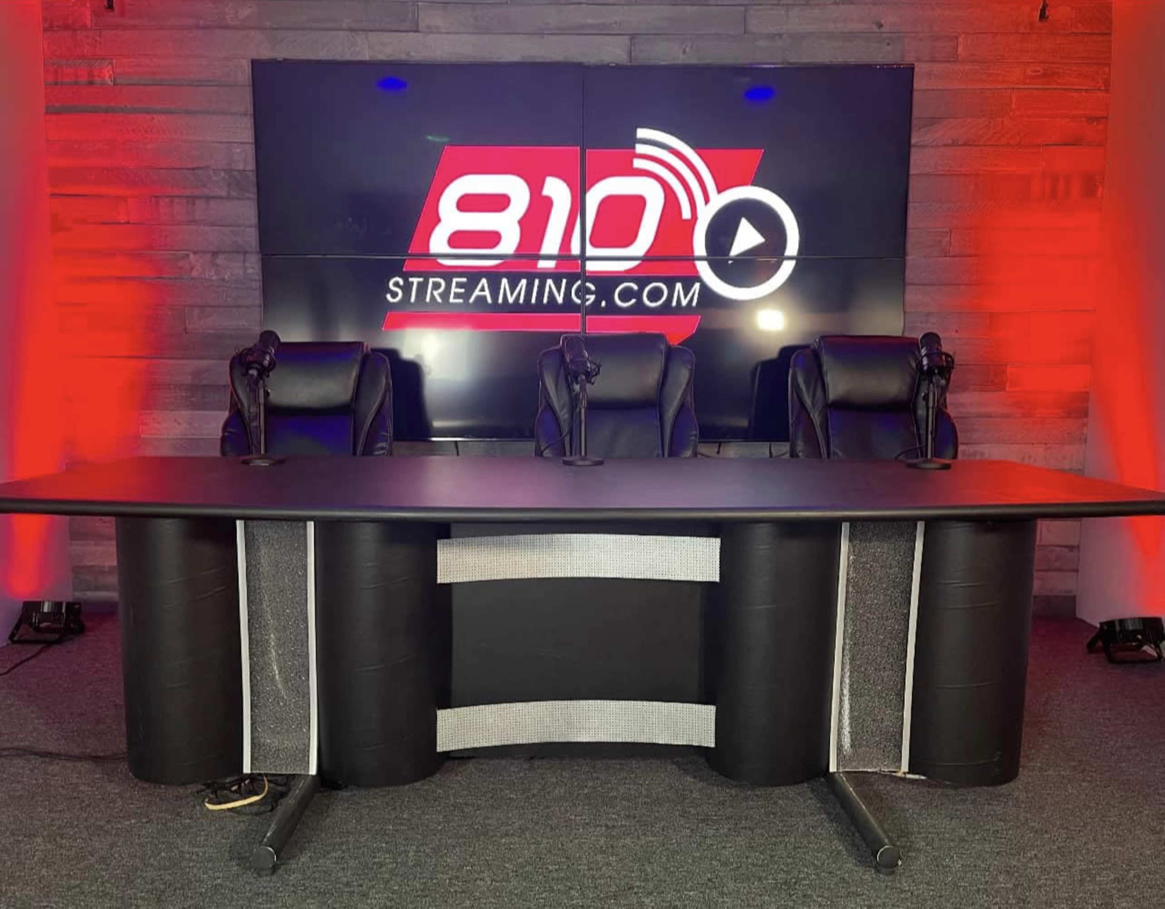 A black table with three empty black chairs sits in front of a large LED screen displaying the text "810 STREAMING.COM."