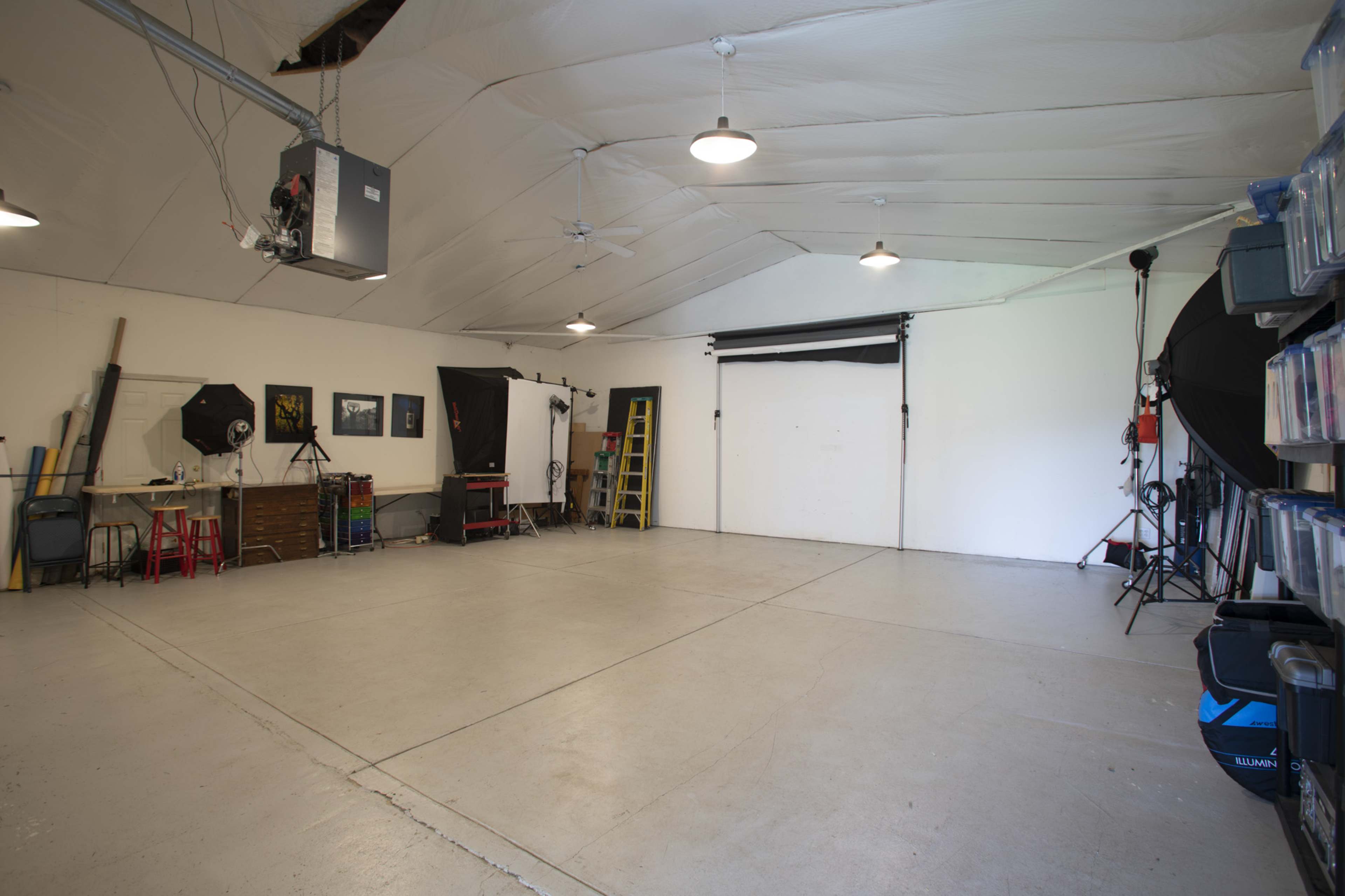 A spacious studio with white walls, a concrete floor, and photography equipment including lights, backdrops, and props organized along one side.