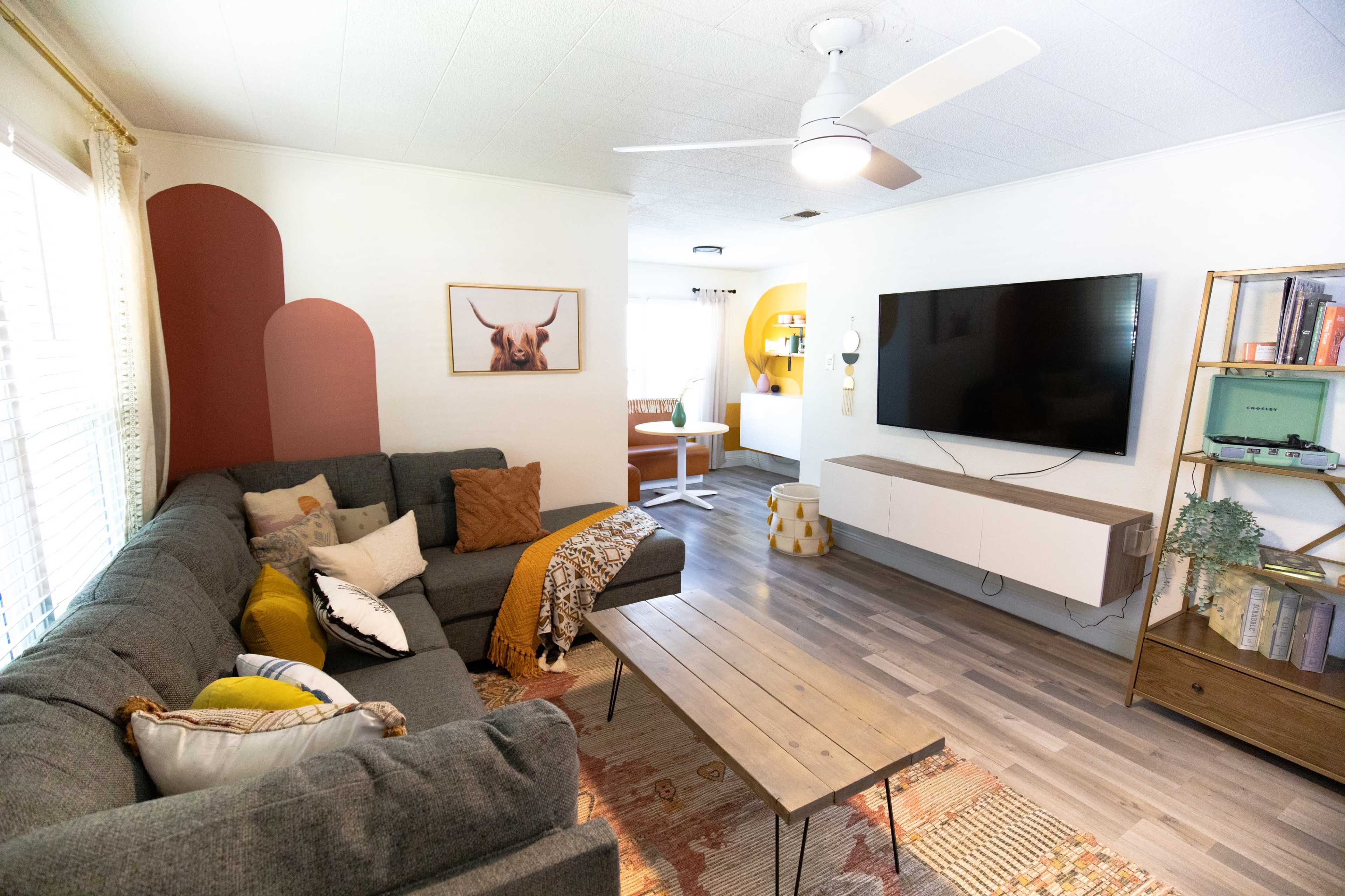 The living room features a sectional gray sofa, a wooden coffee table, a large television mounted on the wall, and decorative elements including plants and artwork.