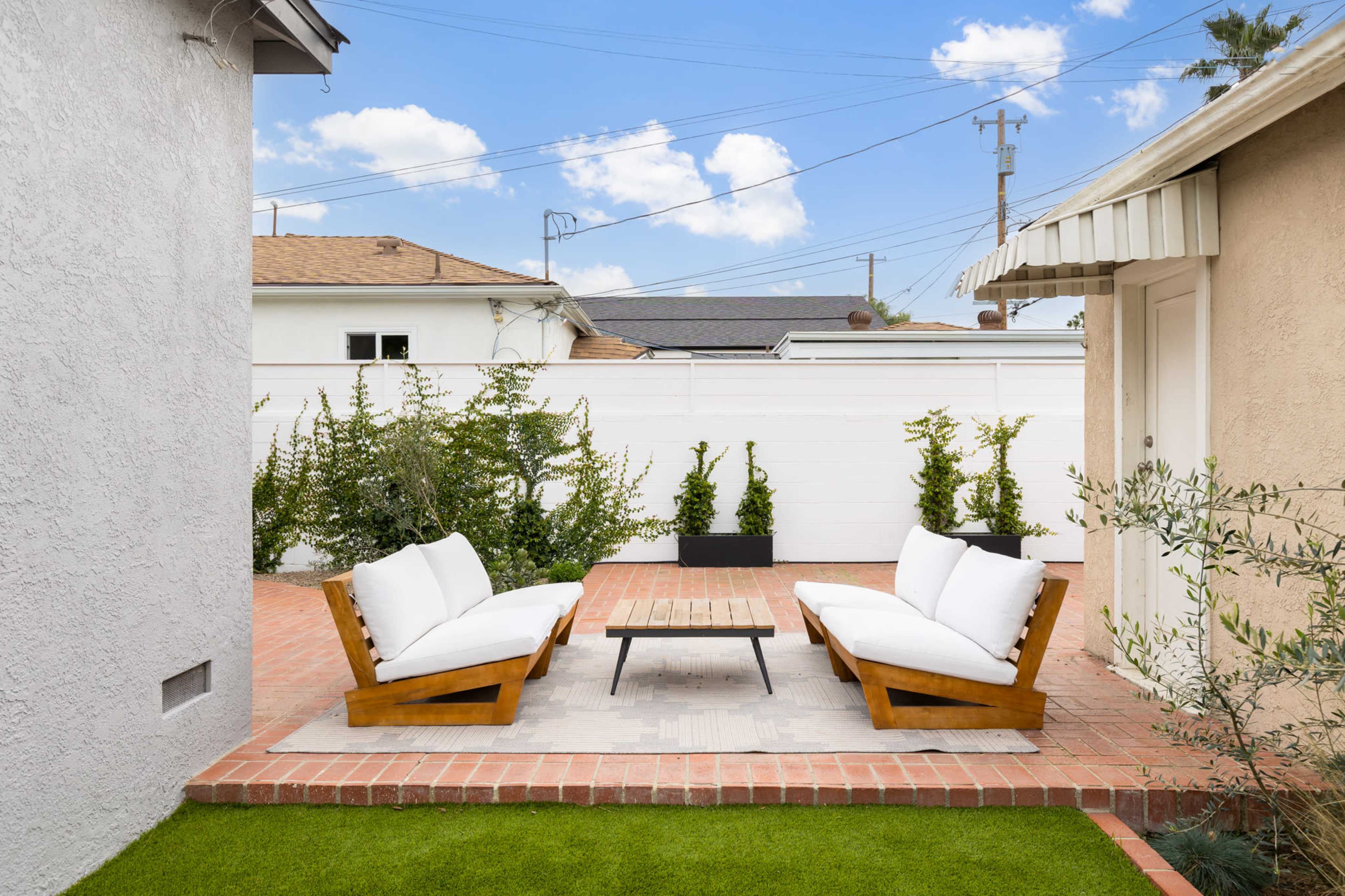 A cozy outdoor seating area features two wooden benches with white cushions arranged around a rectangular coffee table on a brick patio, surrounded by greenery and a white fence.