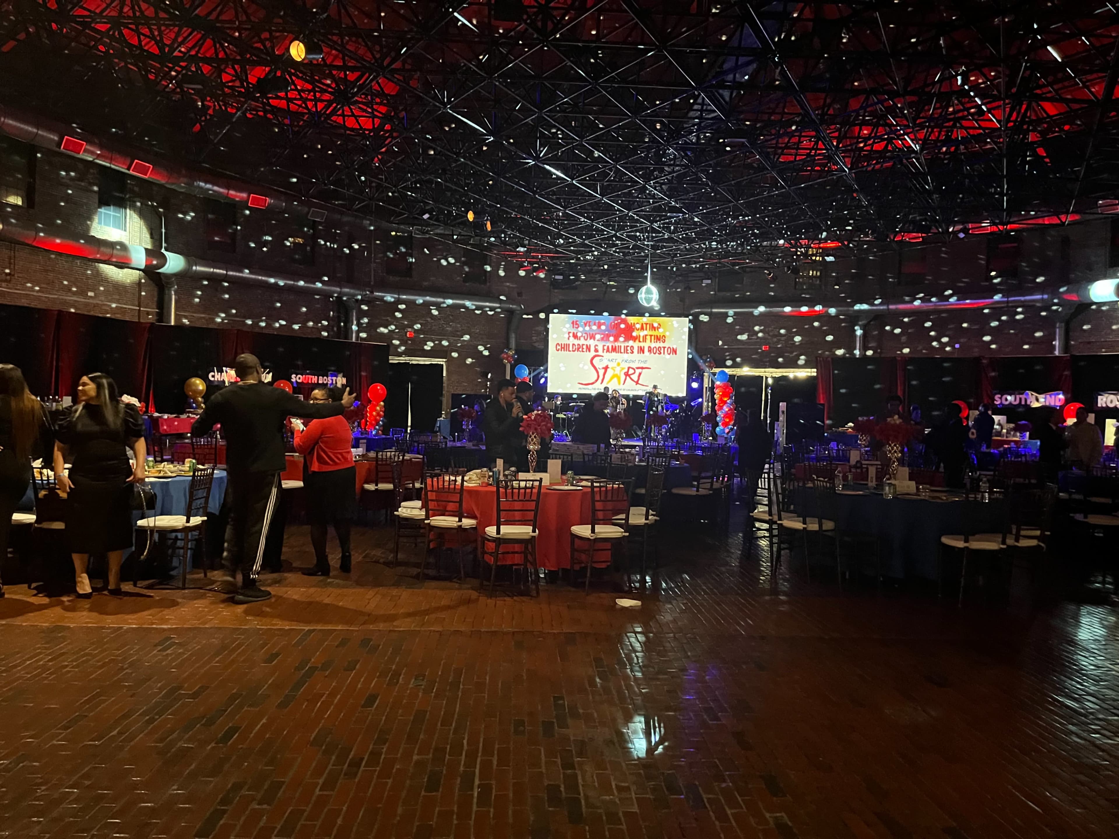 A spacious event hall features decorated tables, colorful lighting, and a large banner that reads "Start" in the background.