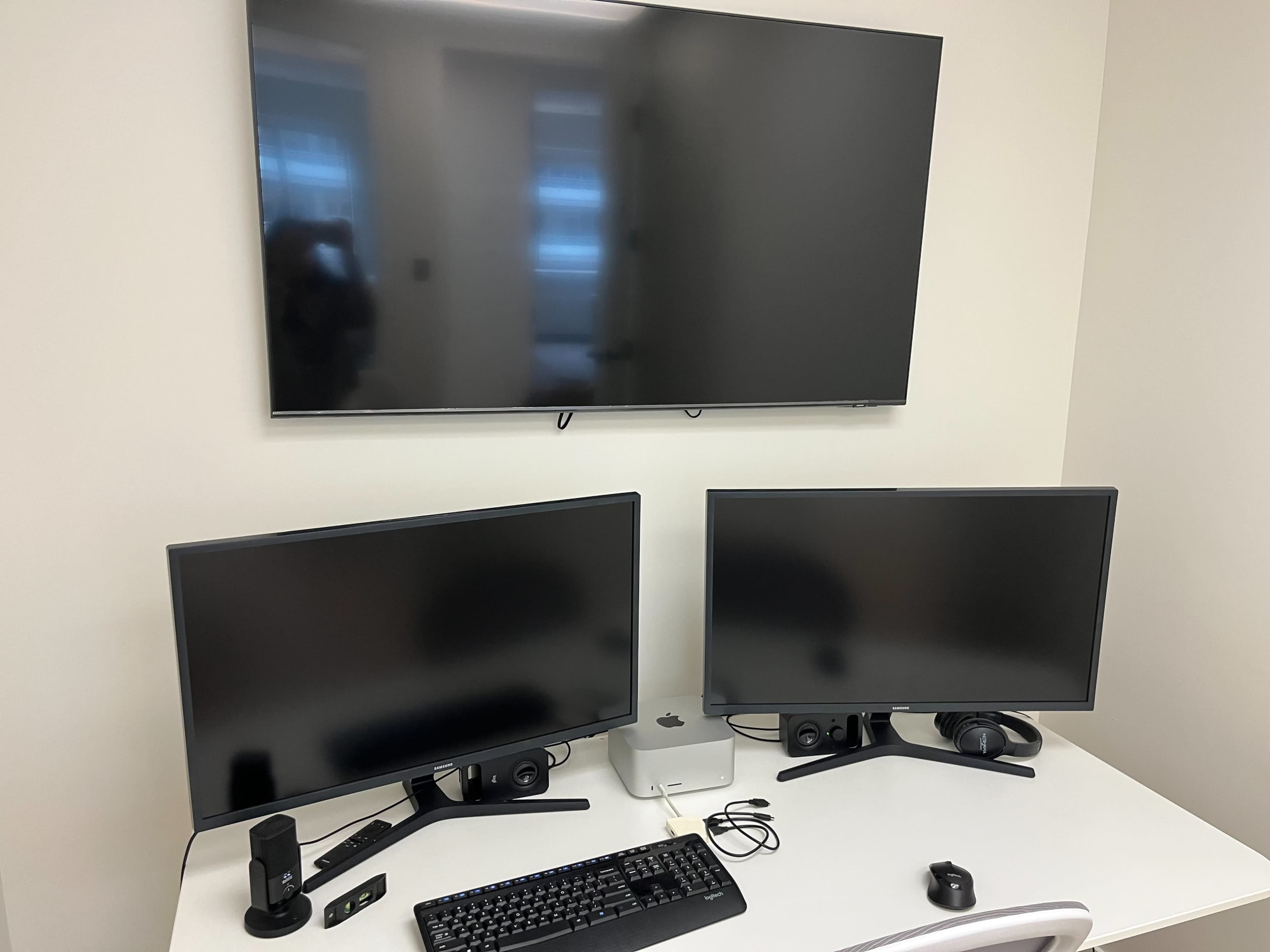 The image shows a workstation with two monitors, a keyboard, speakers, and a webcam on a desk, alongside a large TV mounted on the wall.