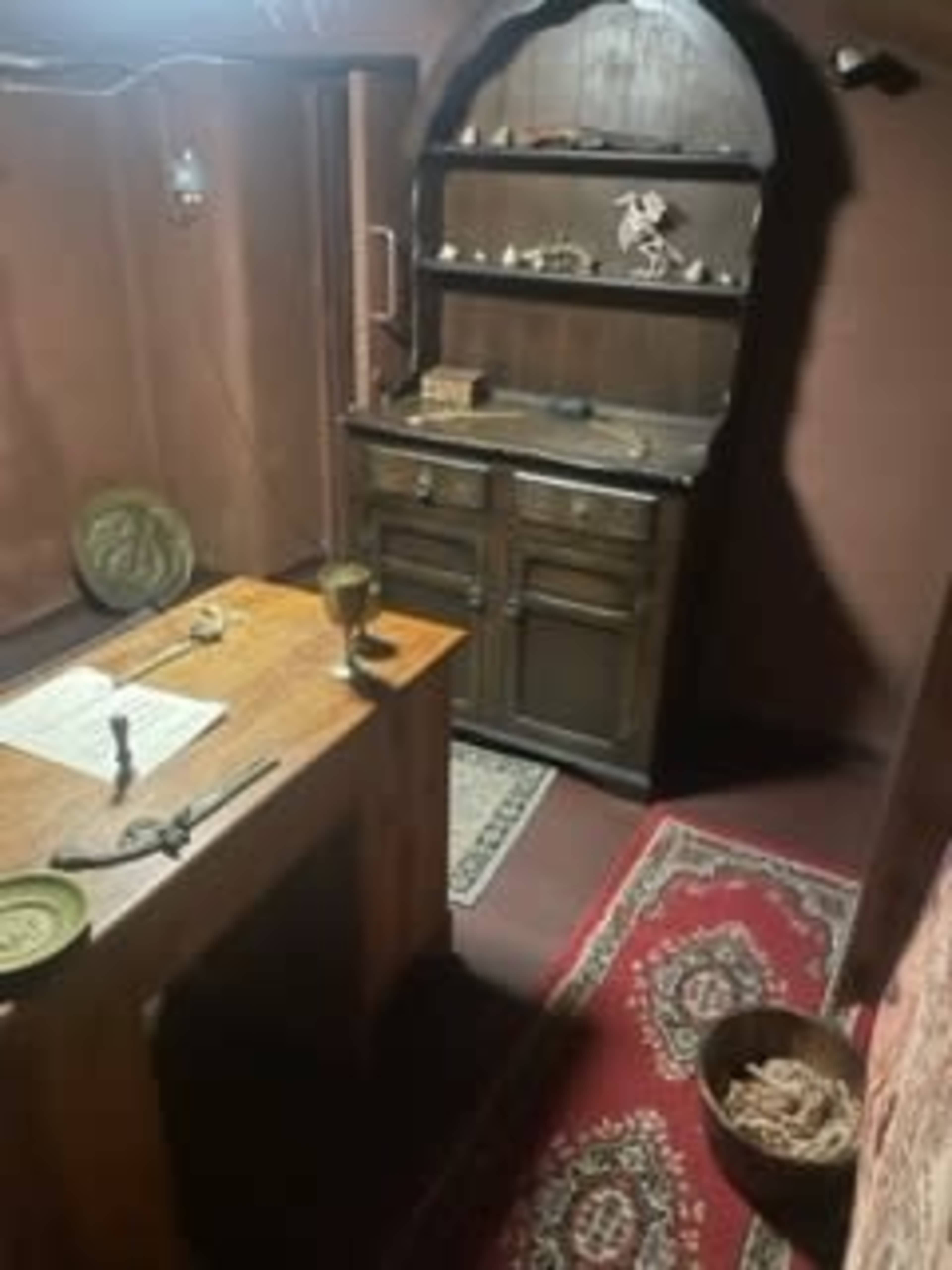 The image shows a dimly lit room featuring a wooden table with an open book, a chalice, and a storage cabinet against the wall, all set on a patterned red rug.