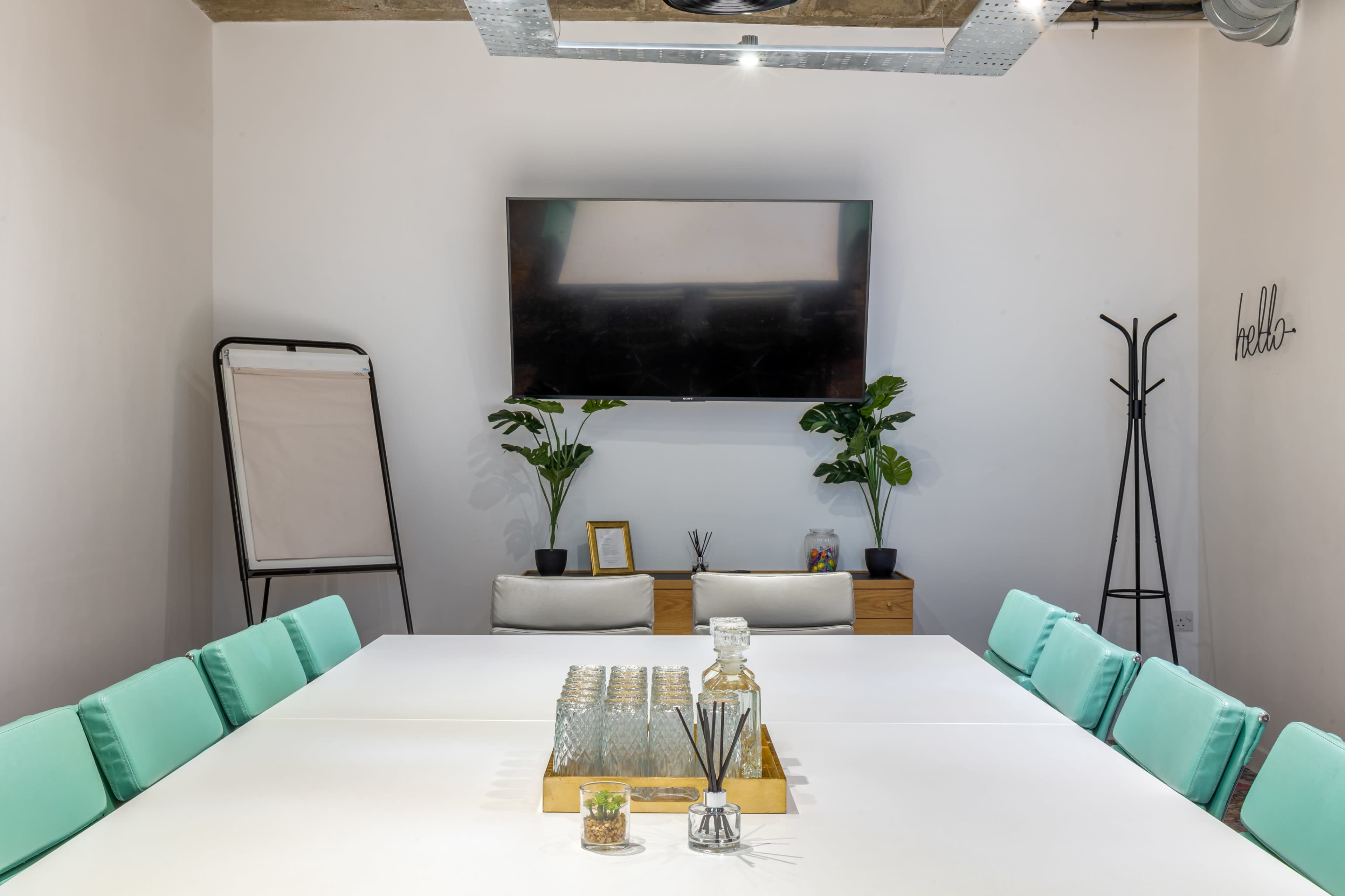 A modern meeting room features a large white table, green chairs, a television mounted on the wall, and potted plants.