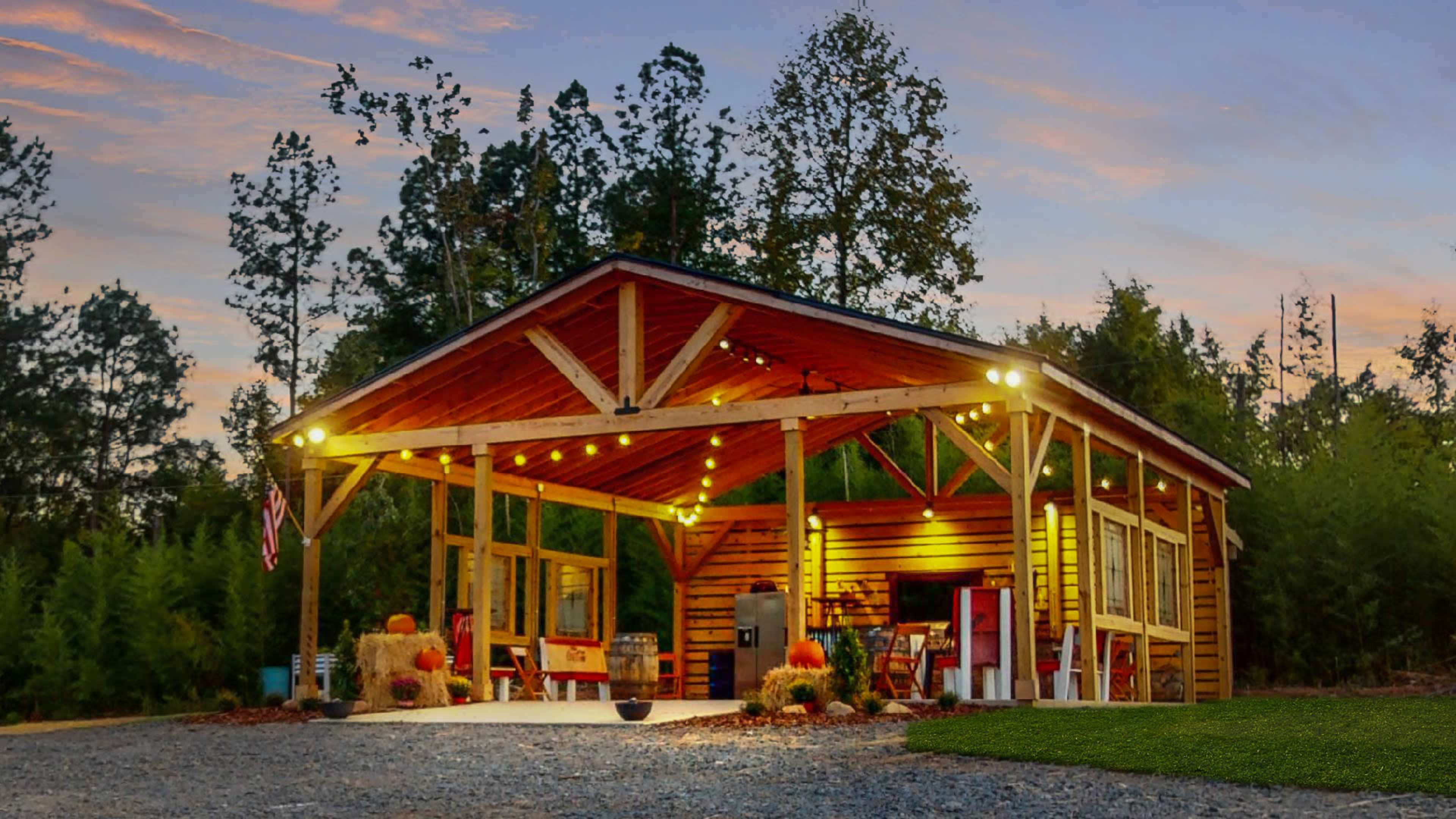 A wooden pavilion with string lights, surrounded by trees, and featuring hay bales and outdoor seating.