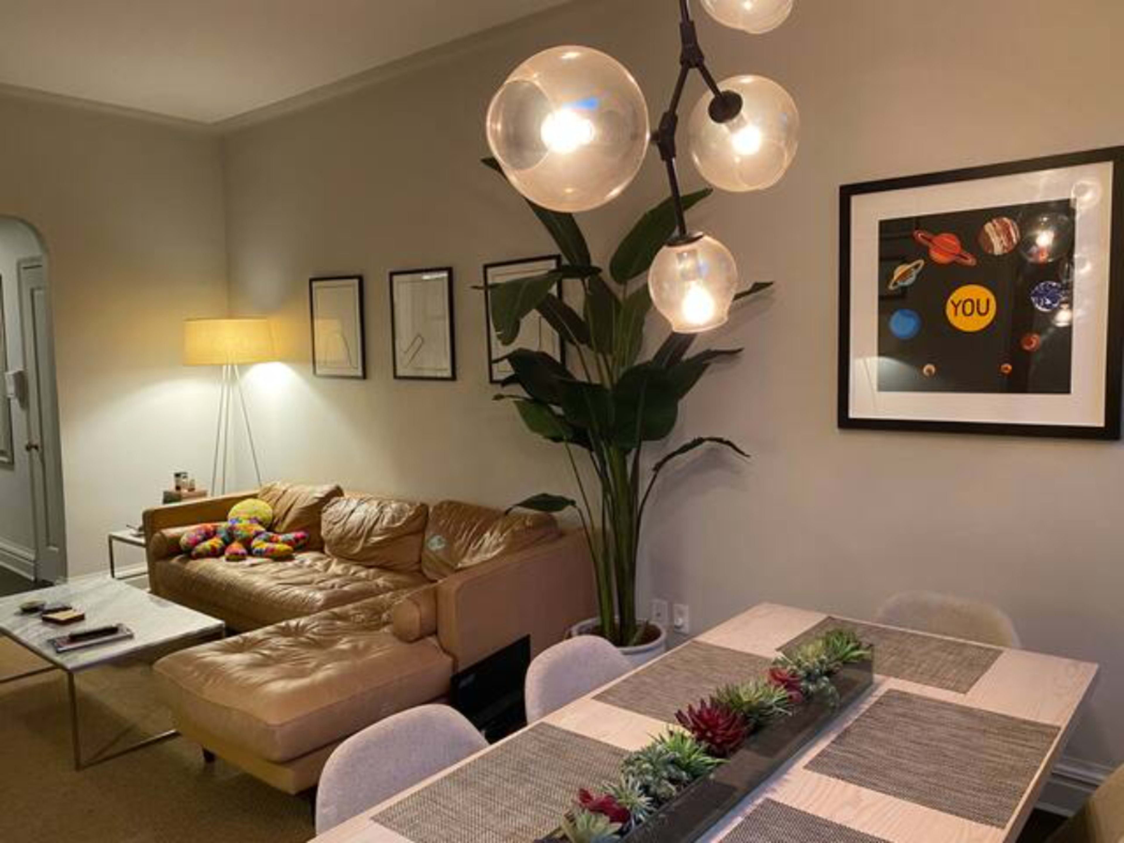 A living and dining area featuring a beige leather sofa, a large framed artwork on the wall, modern light fixtures, and a dining table with decorative plants.