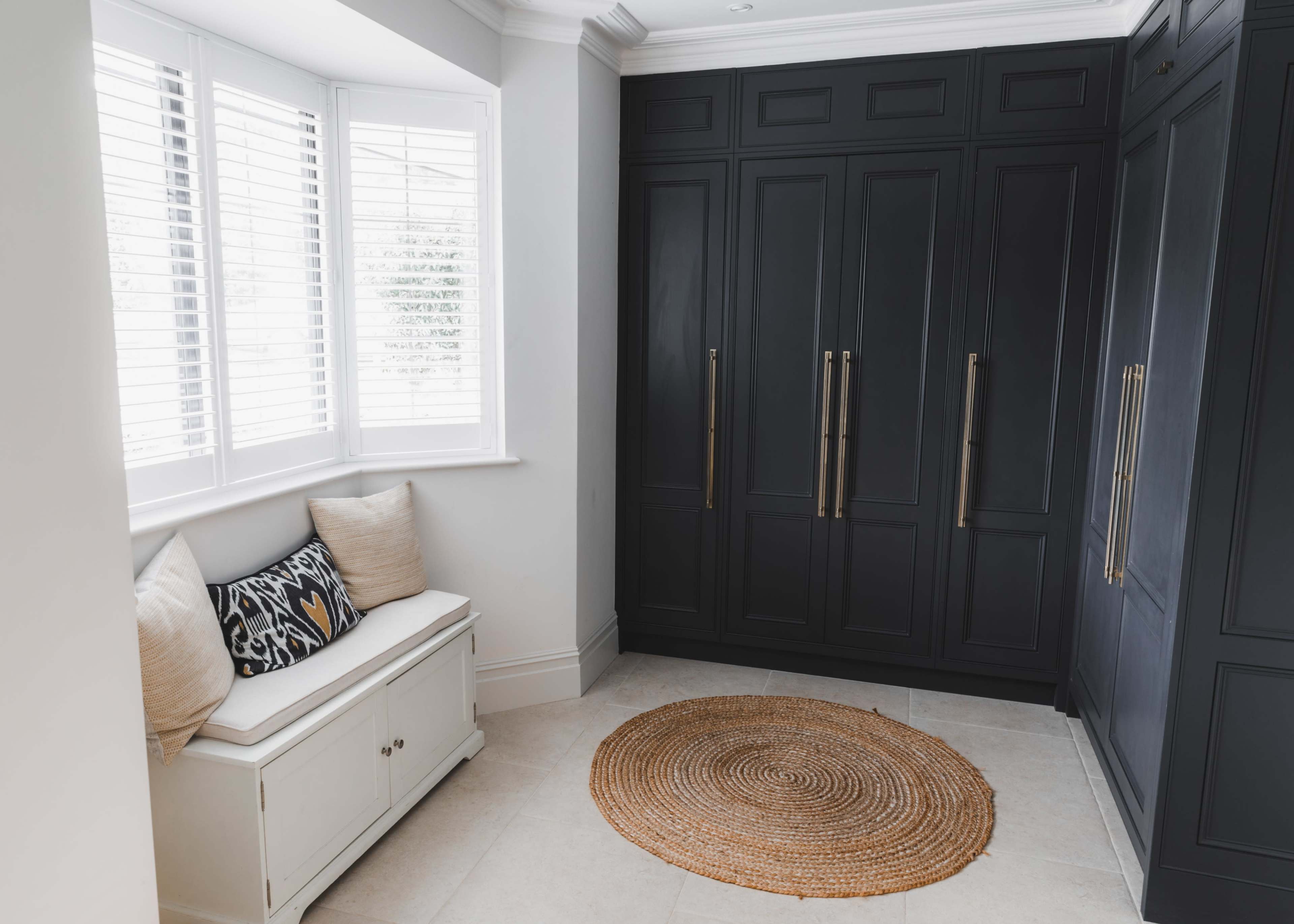 A hallway with a dark, built-in wardrobe, a small white bench with decorative cushions, and a round, woven rug on a tiled floor.