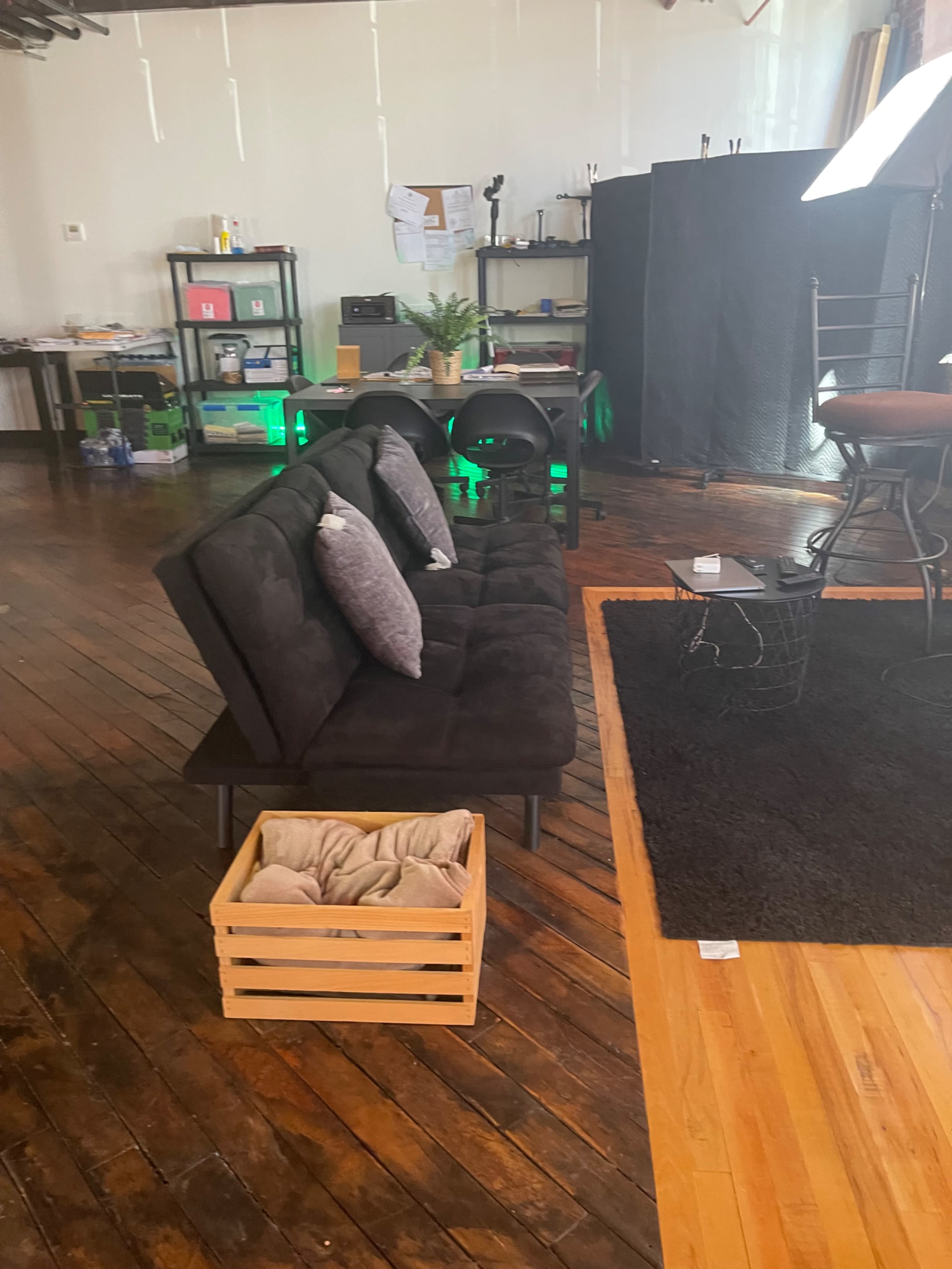 A modern black sofa with cushions, a wooden crate with a blanket, and a rug on a hardwood floor in a room filled with various furniture and equipment.