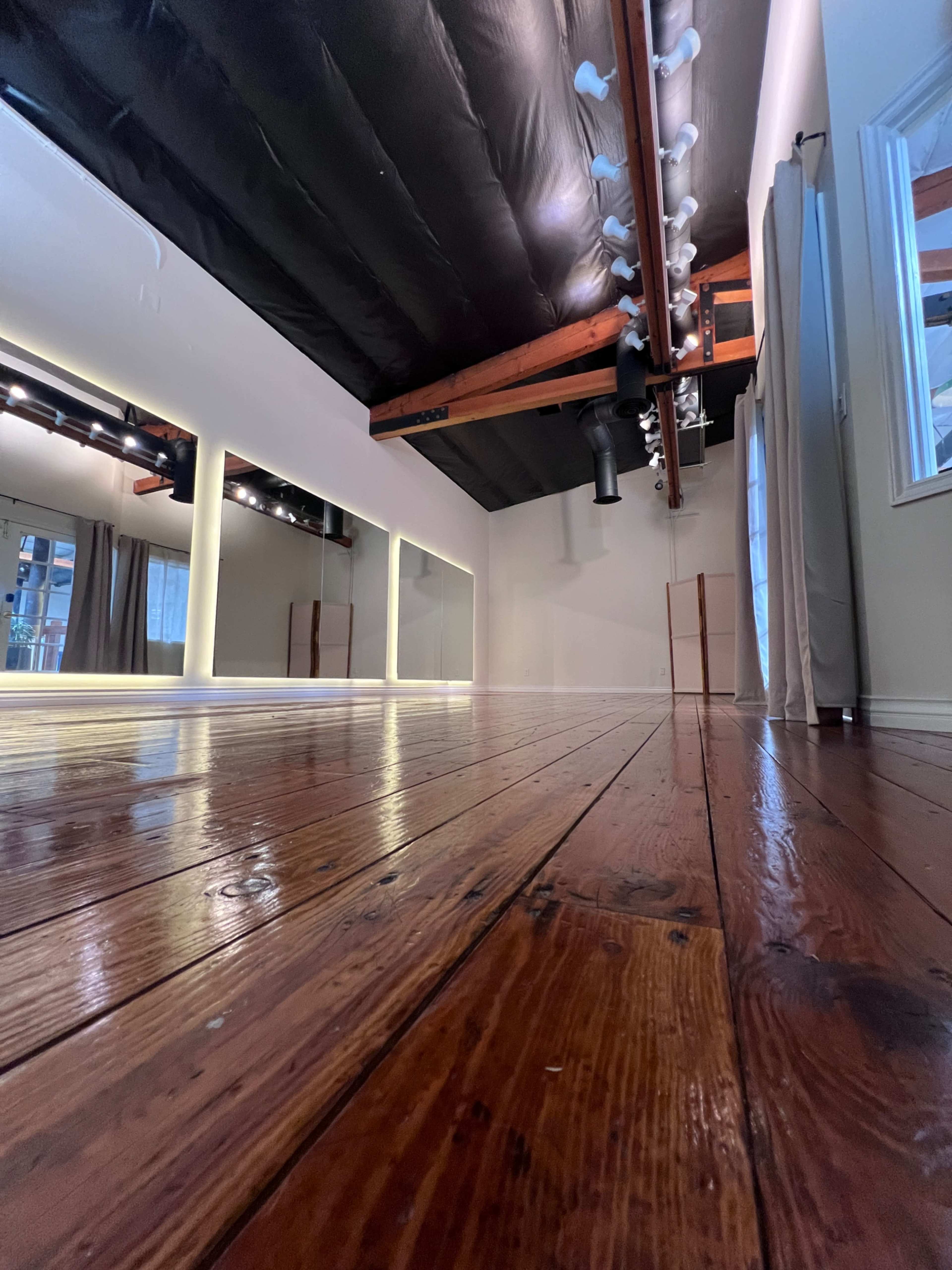 The image shows an empty dance studio with wooden flooring, mirrors along the wall, and soft lighting.