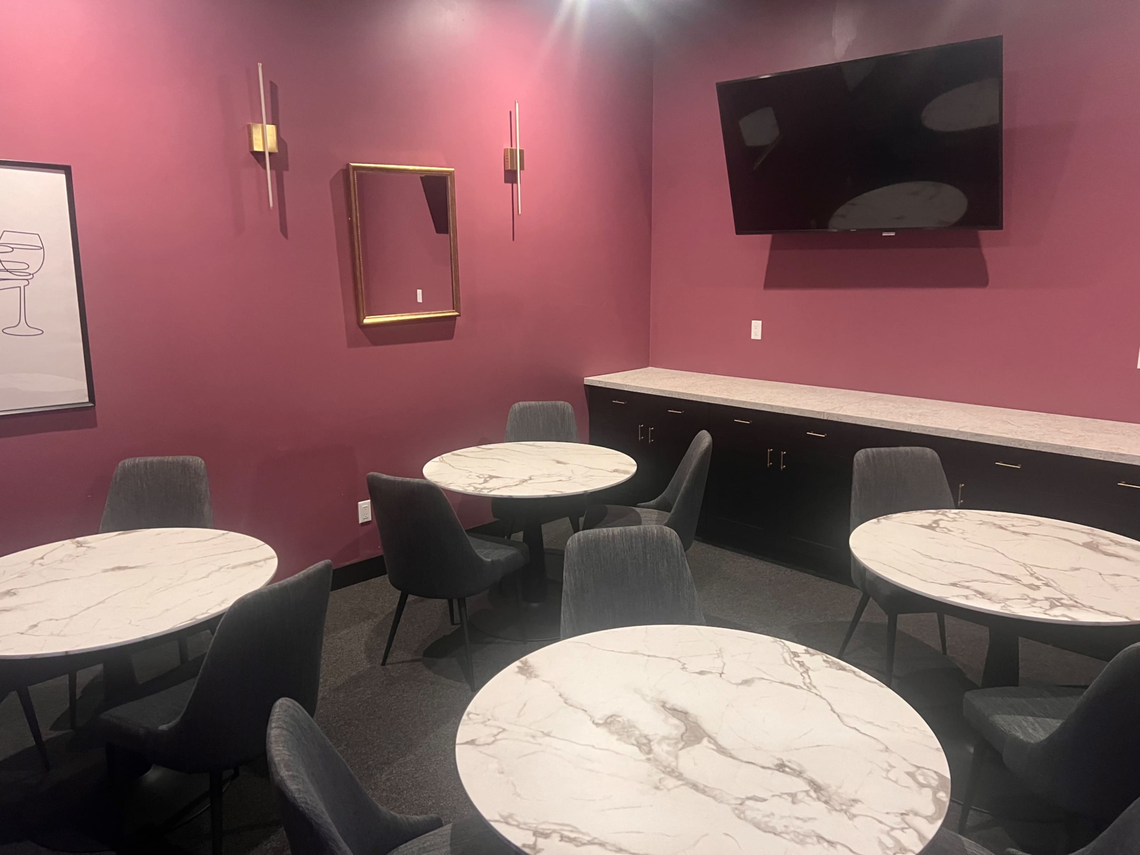 A small, modern dining area with round marble tables and black chairs against a deep pink wall, featuring a mounted television and a mirror.