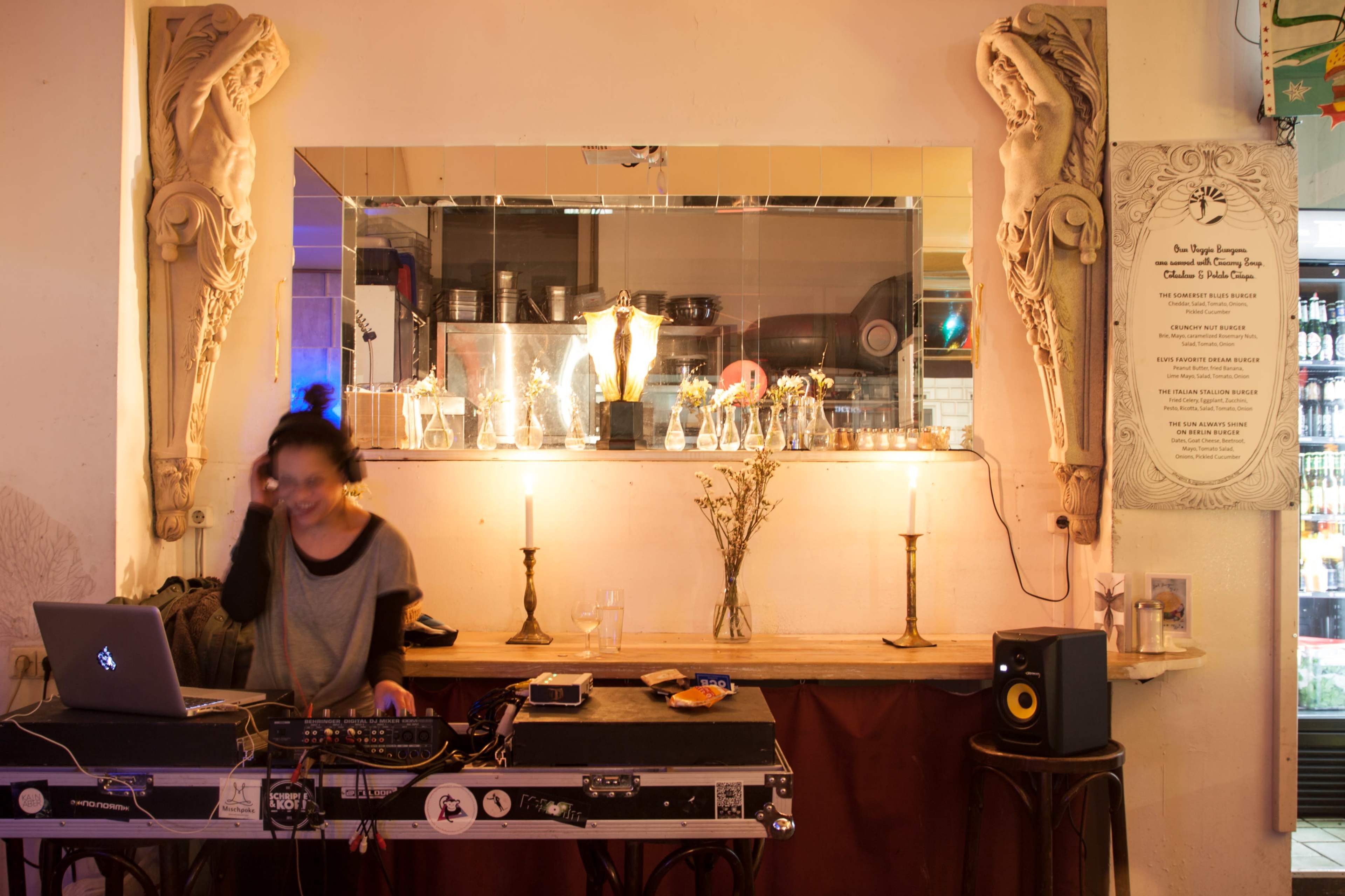 A DJ stands behind a table with equipment in a dimly lit bar, featuring decorative wall carvings and illuminated candles on a counter.