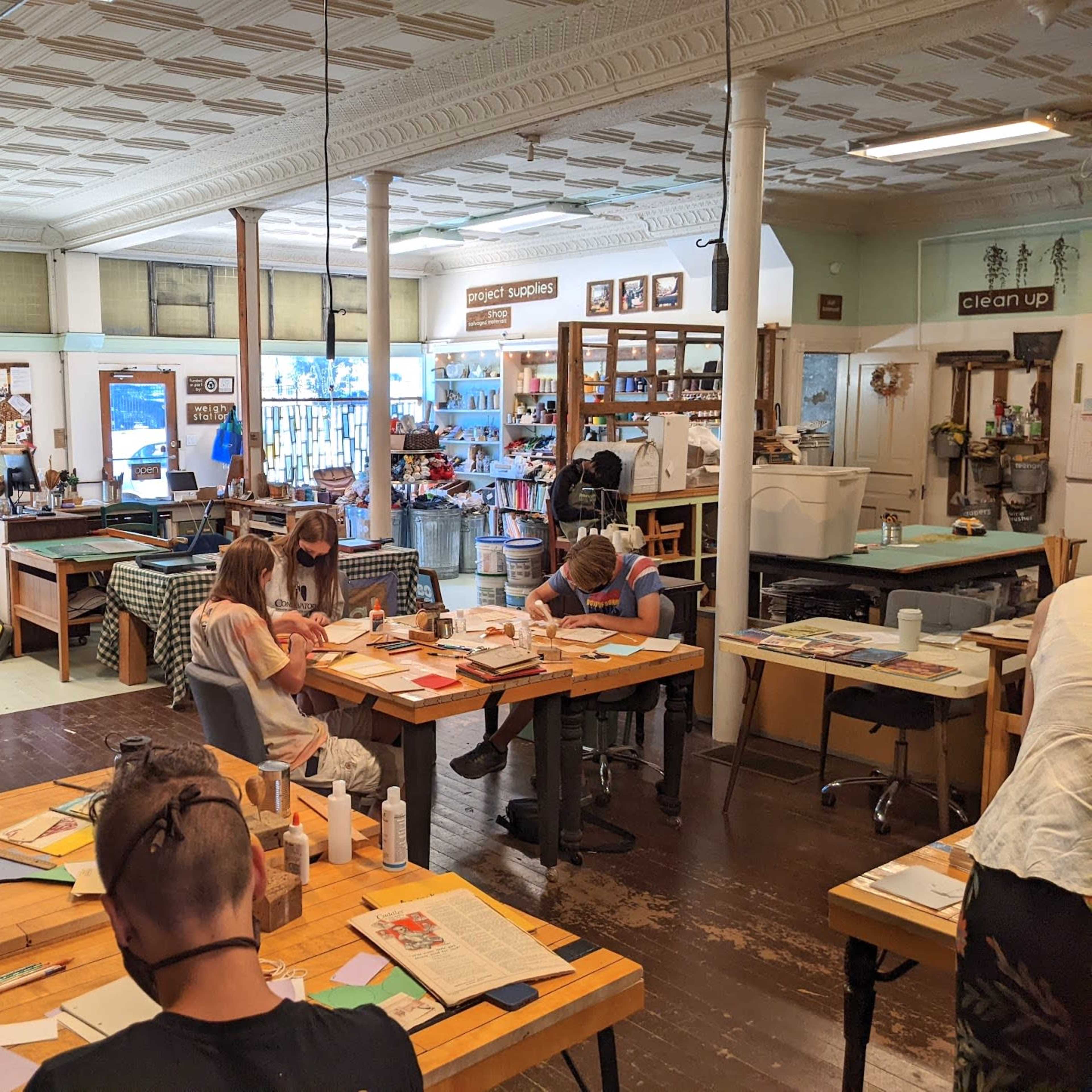 A group of people are seated at wooden tables in a creative workshop, engaged in various art projects.