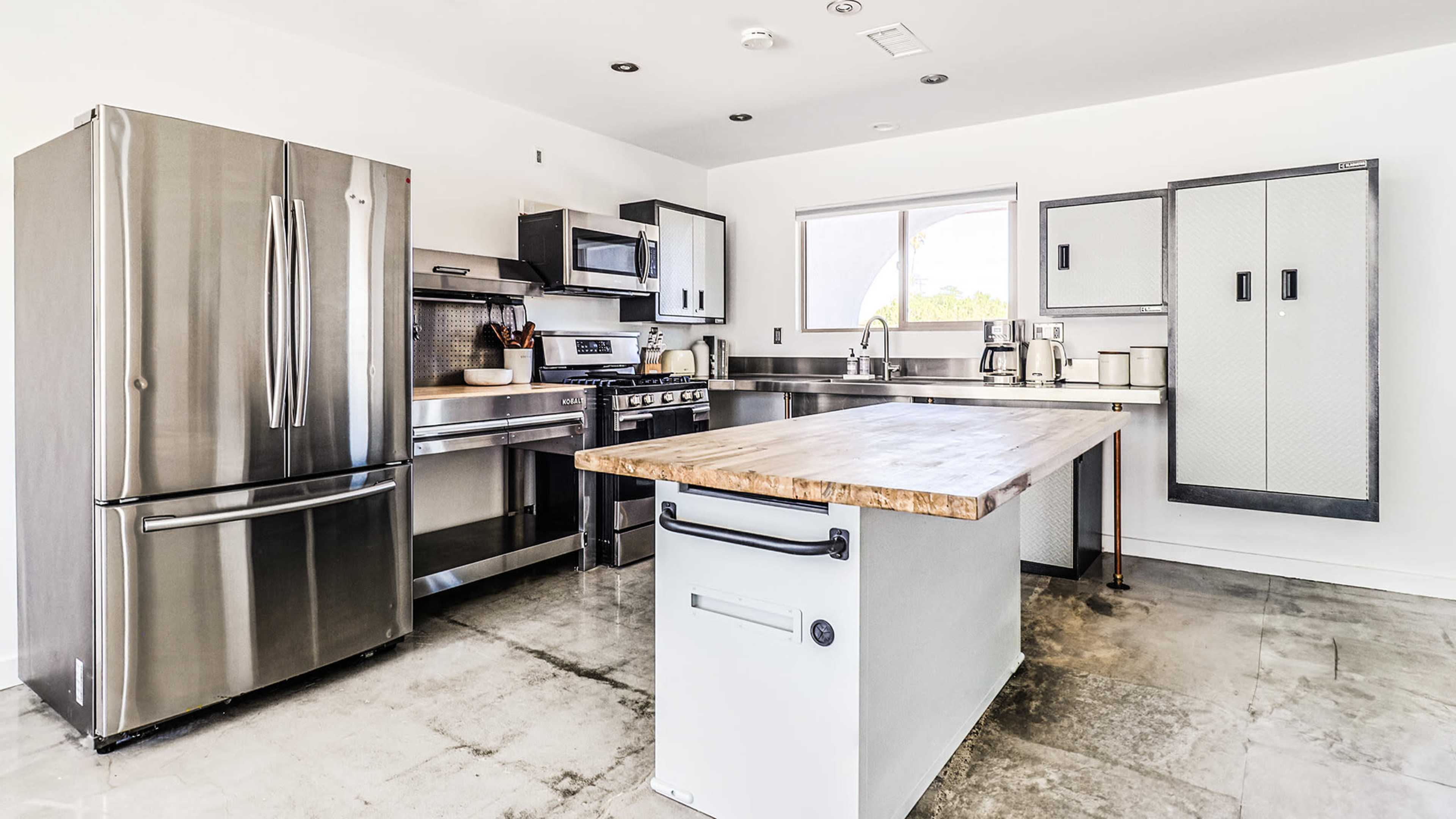 A modern kitchen featuring stainless steel appliances, a central wooden island, and minimalist cabinetry against a white wall.