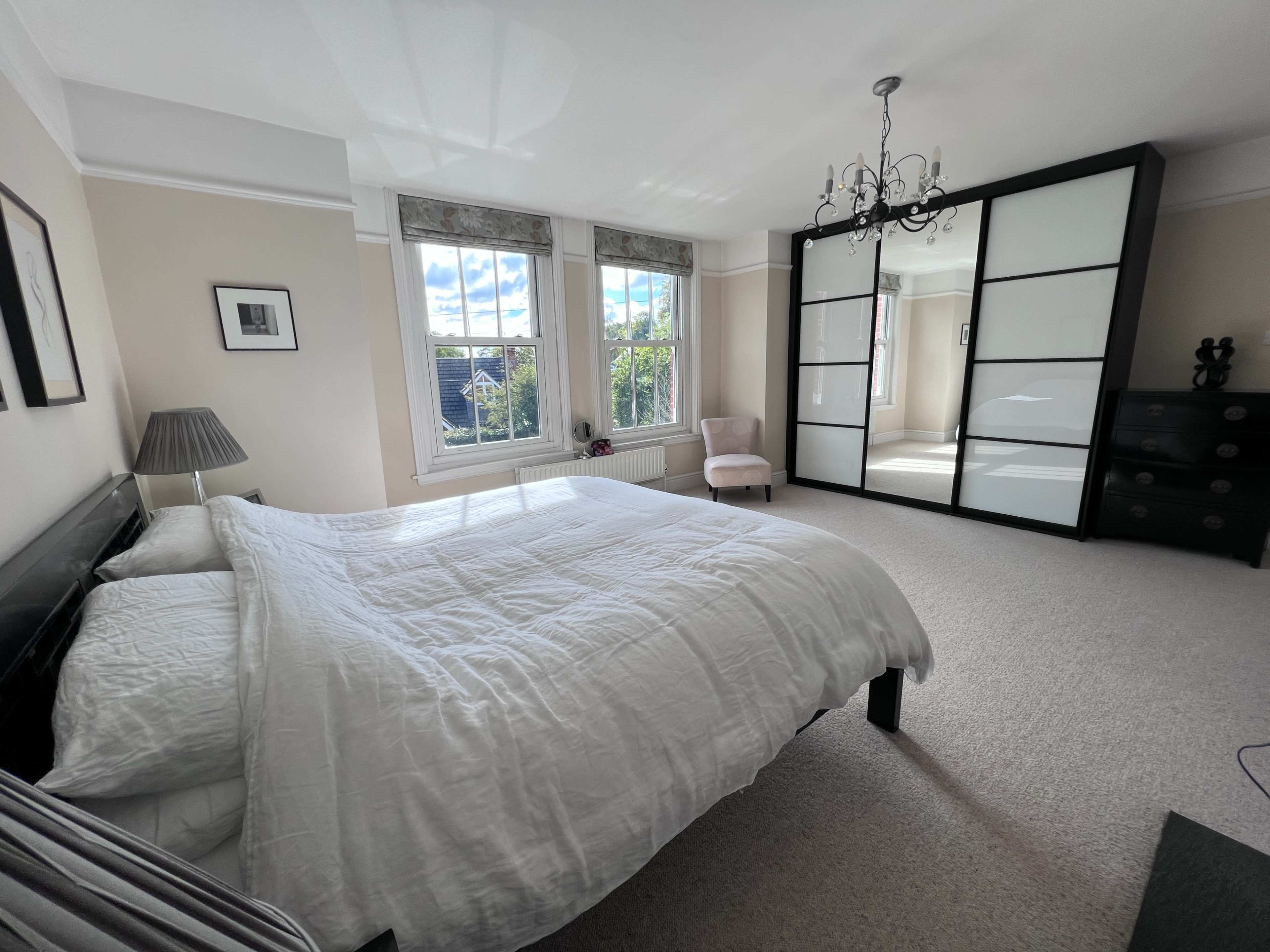 A bright, spacious bedroom features a large bed, a chandelier, a seating area, and a wardrobe with mirrored doors.