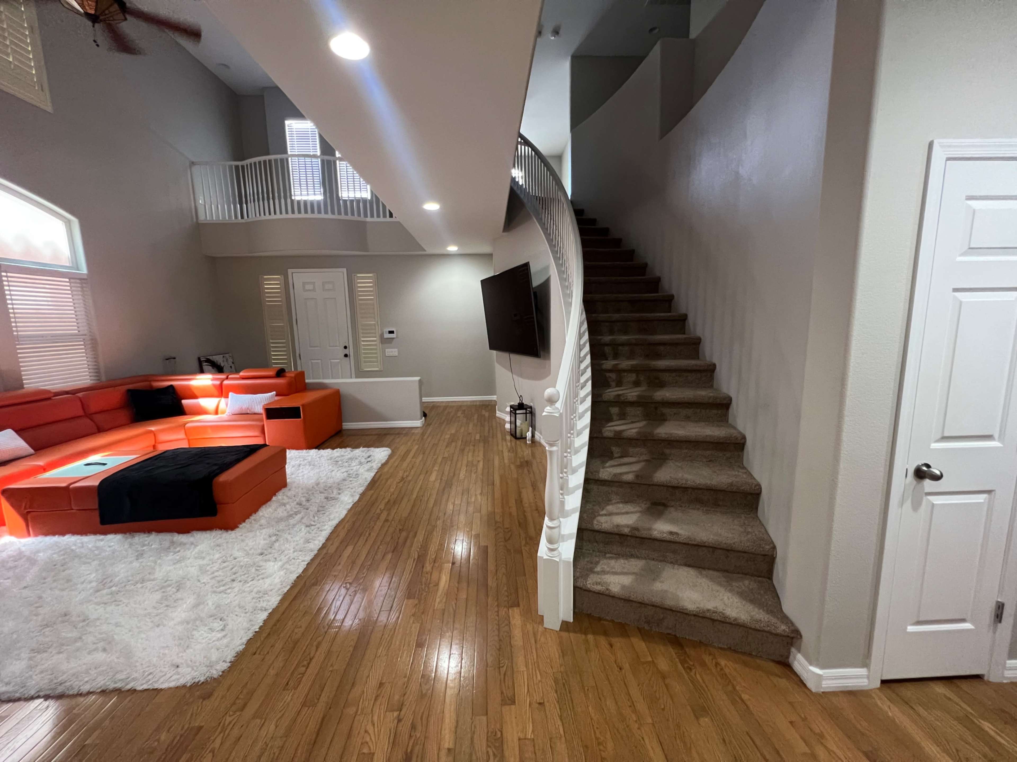 A spacious living area with an orange sectional couch, a large white rug, and a staircase leading to an upper level.