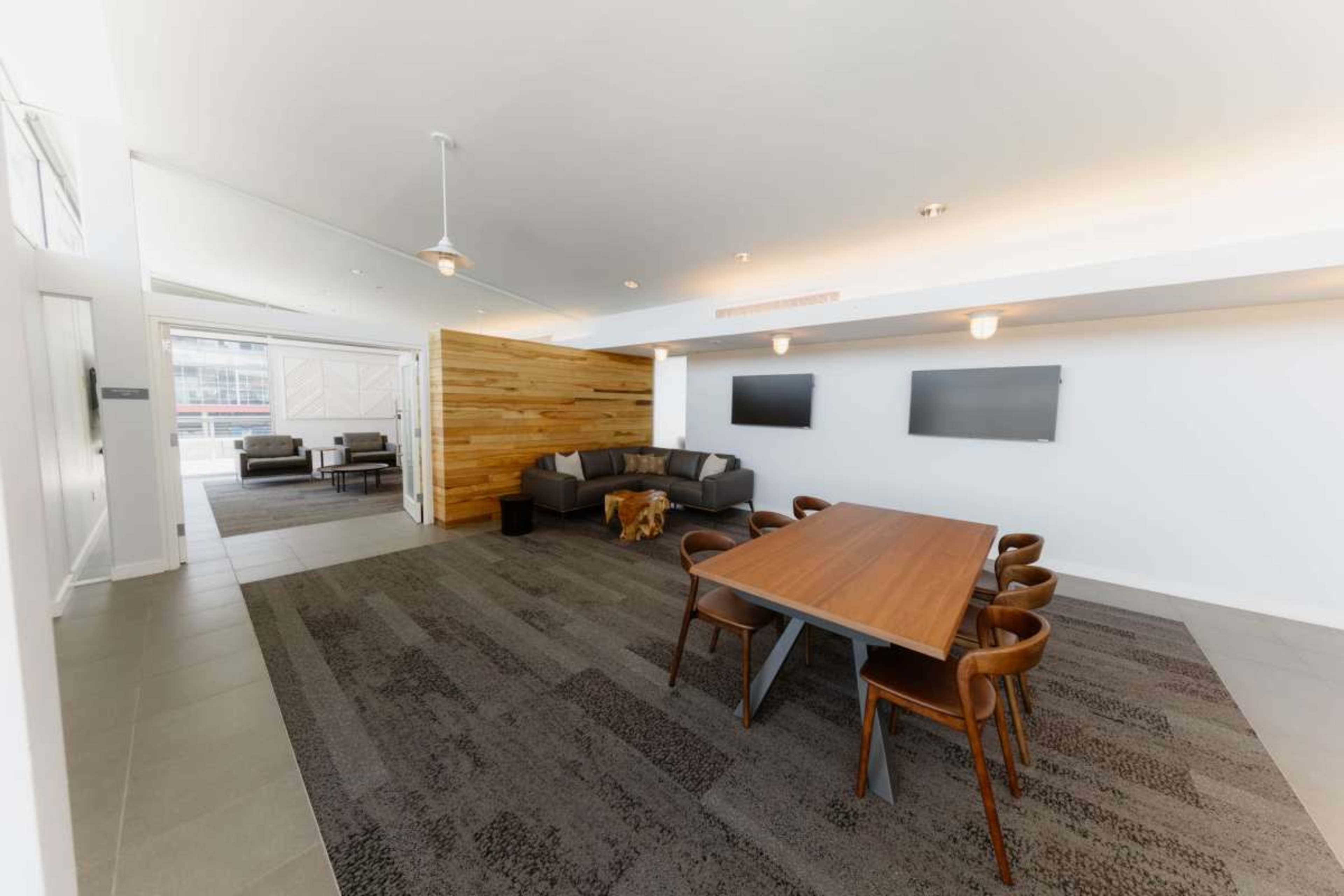 The image shows a modern, open space with a dining table surrounded by chairs, a seating area with couches, and two wall-mounted televisions.