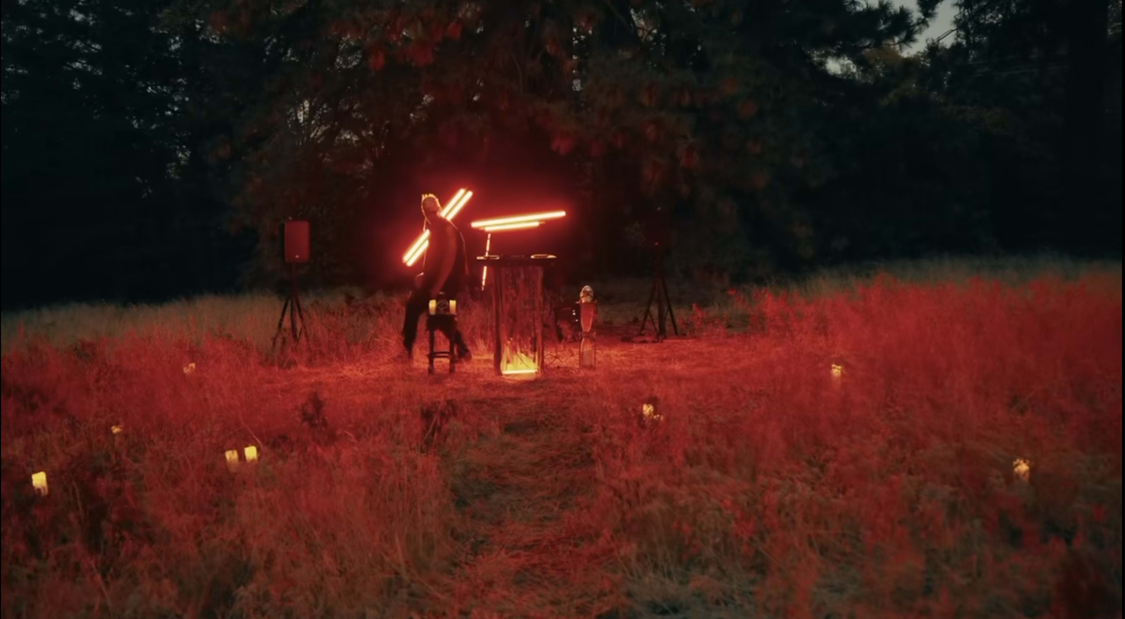 A performer stands in an outdoor field illuminated by red lights, with speakers and candles arranged on the ground.