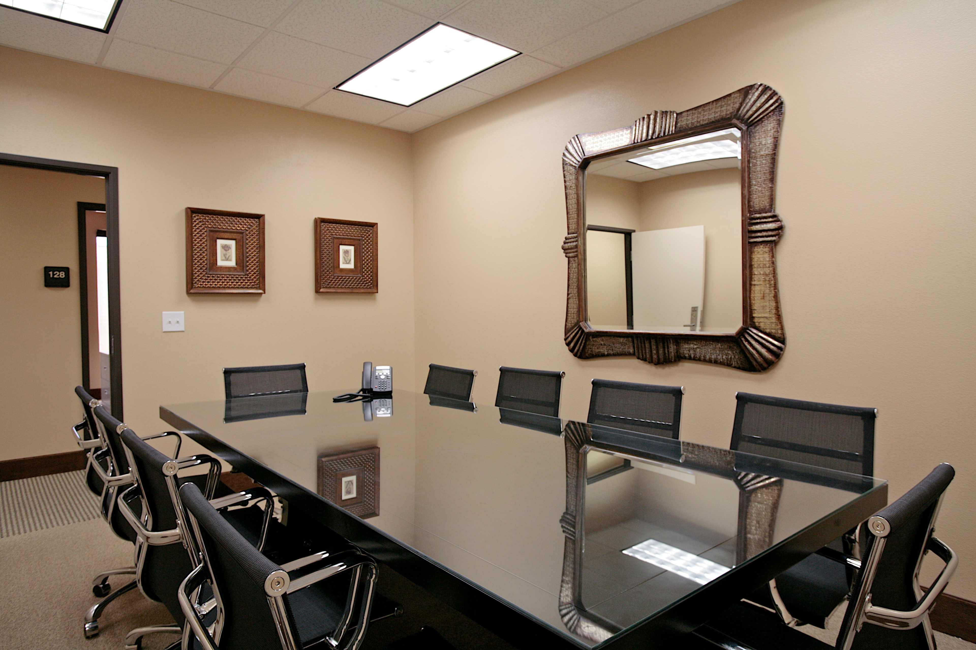 A conference room features a long glass table surrounded by black chairs and is accented by two framed artworks and a decorative mirror on the wall.