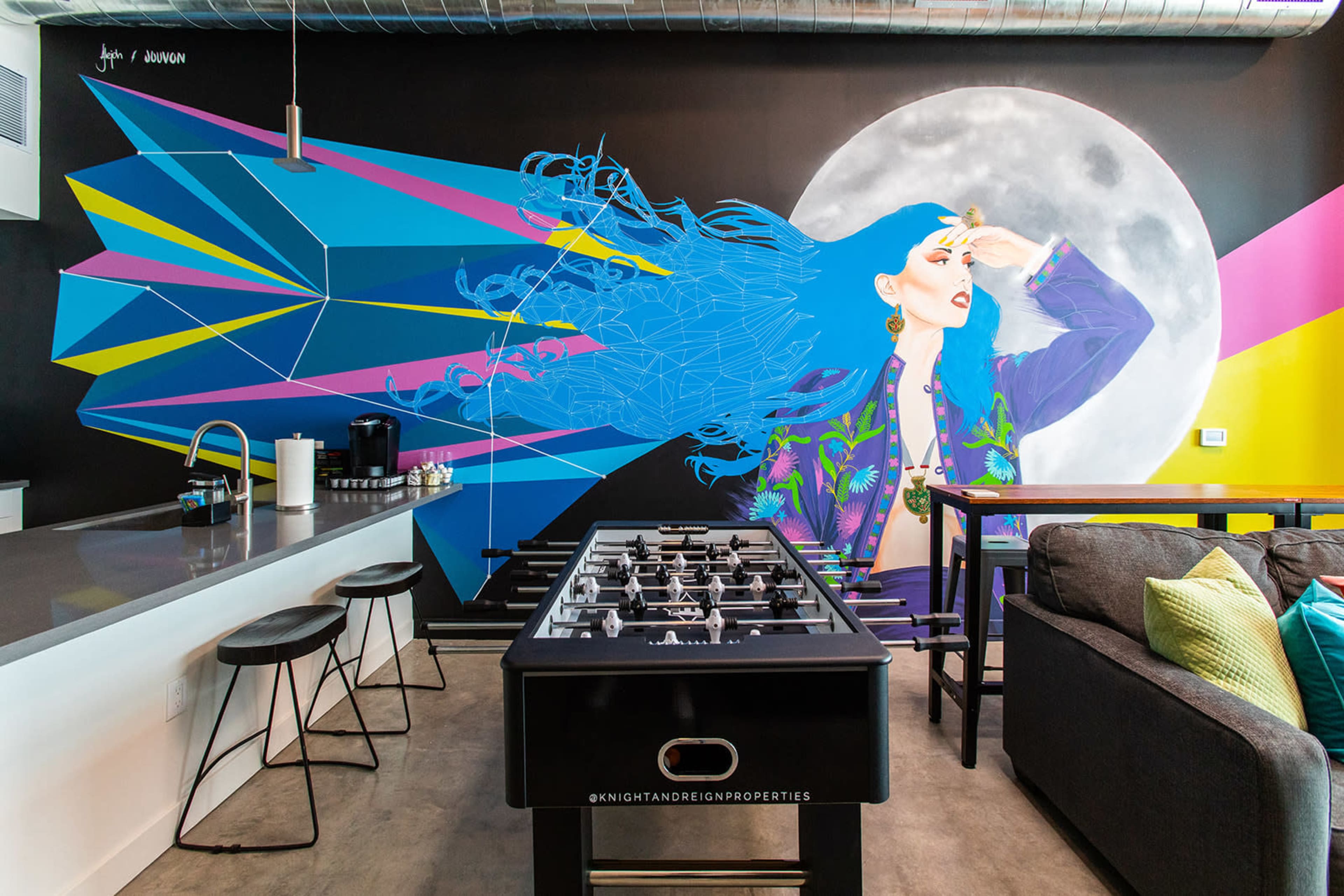 A modern interior with a foosball table in the foreground and a vibrant mural of a woman against a geometric background on the wall behind a kitchen area.