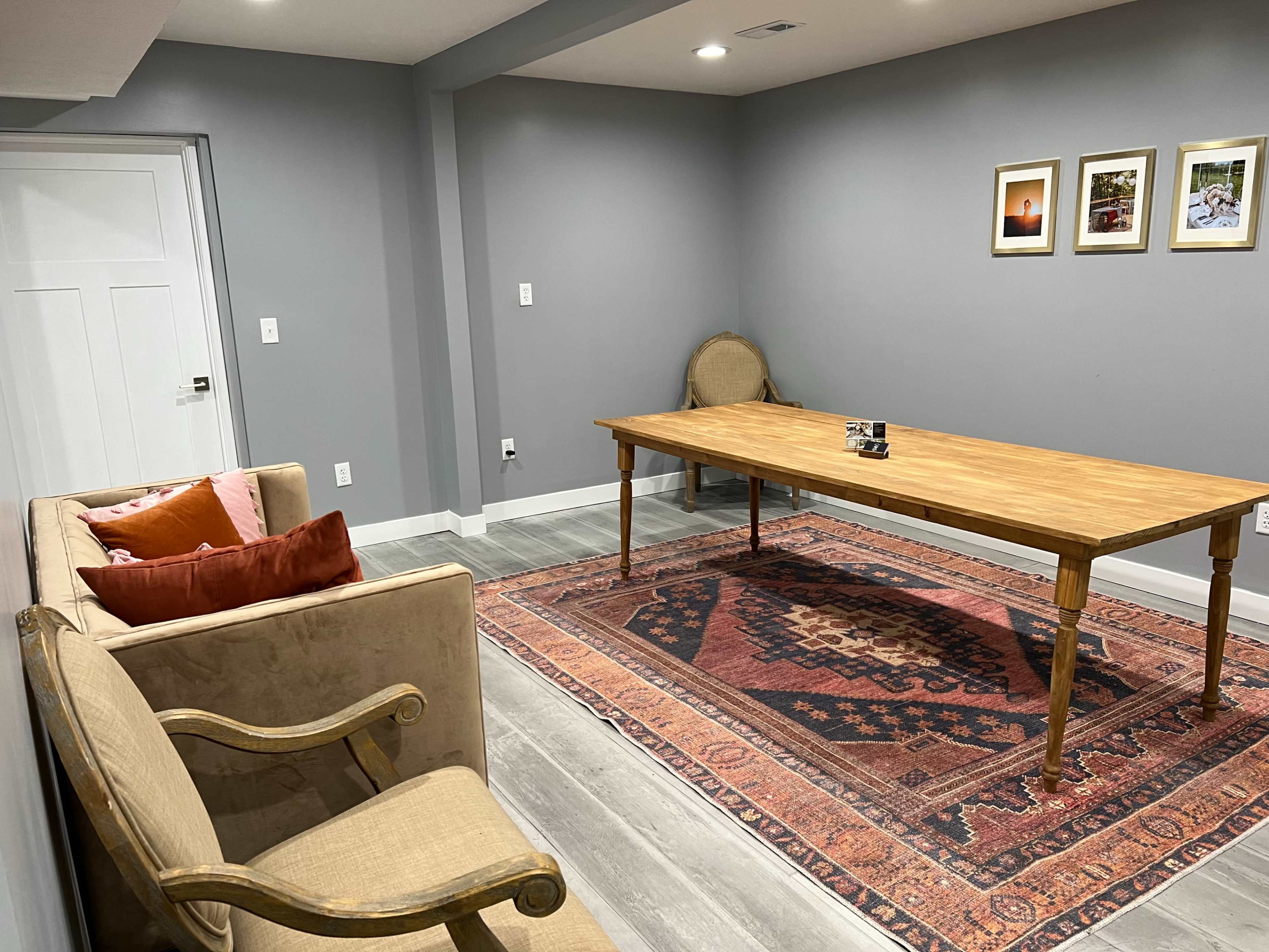 A simple interior room with a wooden table, two chairs, a couch with cushions, and framed pictures on the gray wall.