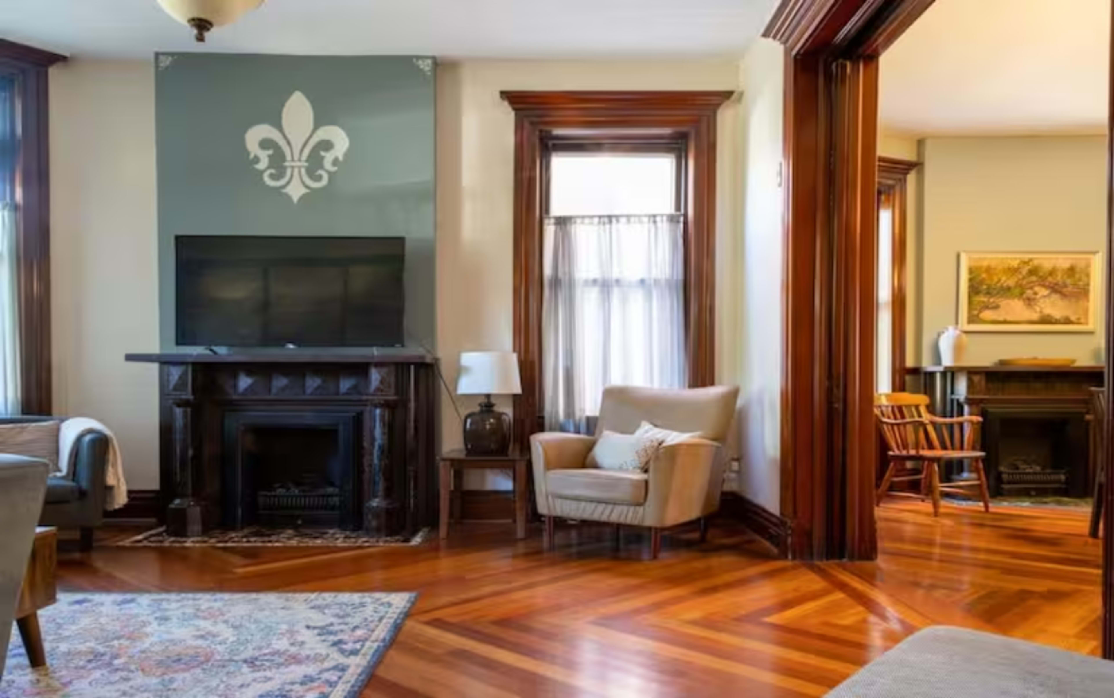 The image shows a well-furnished living room featuring a television above a fireplace and a cozy seating arrangement with large windows.