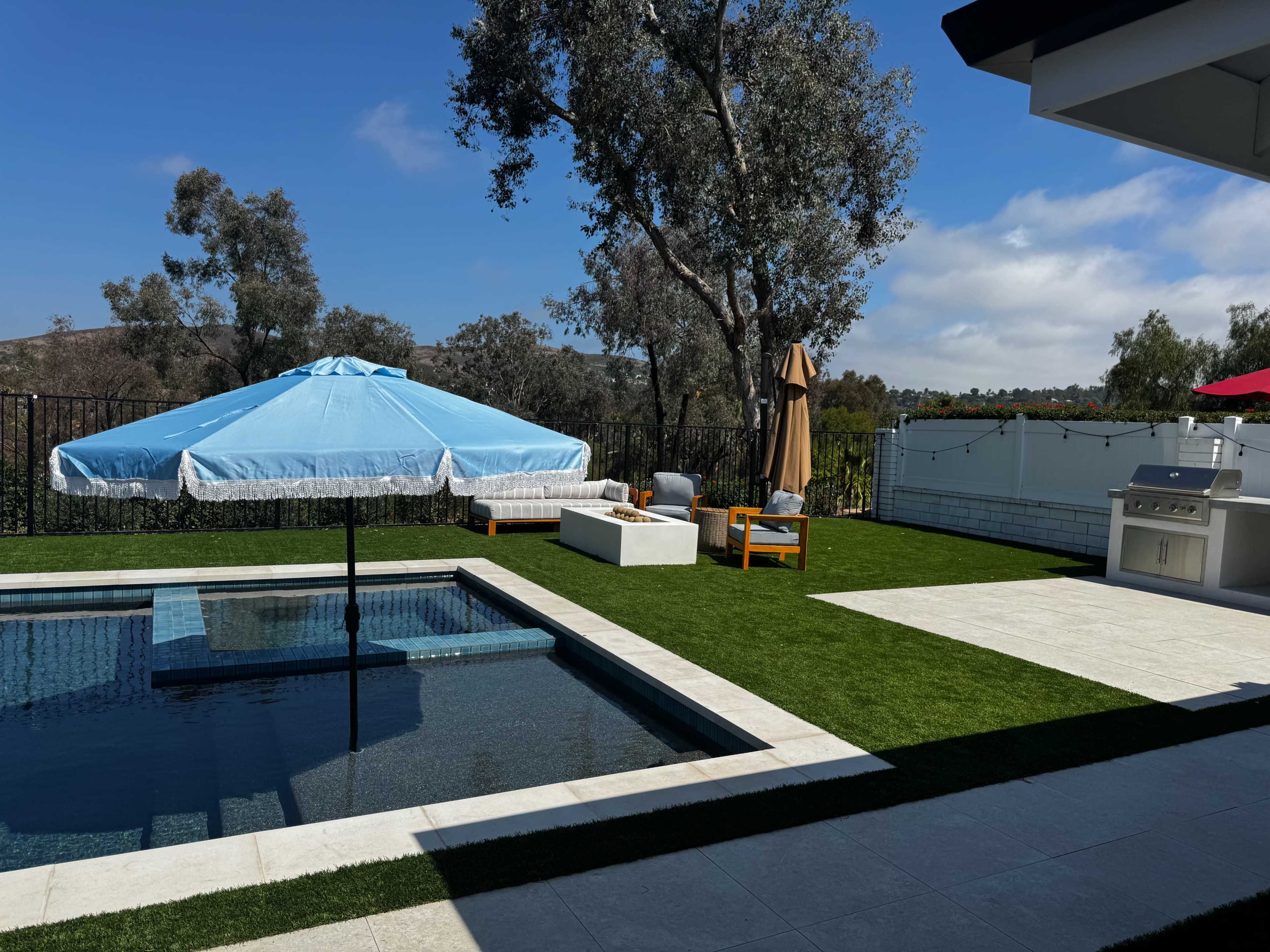 The image shows a backyard with a swimming pool, a large blue umbrella, a white seating area, and a barbecue grill near an outdoor kitchen, all surrounded by grass and trees.