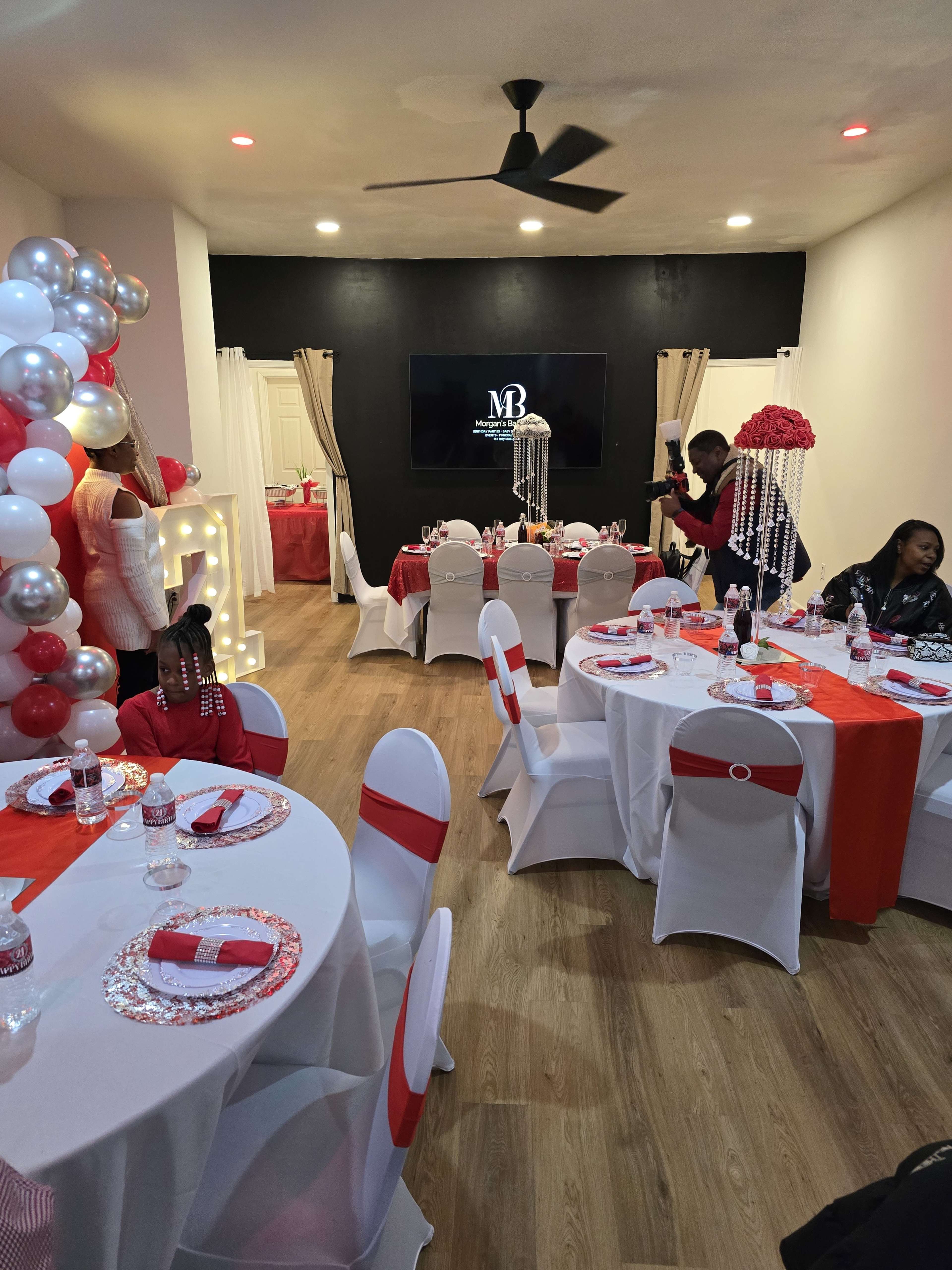 The interior of a banquet room is decorated with red and silver accents, featuring round tables set with plates and water bottles, while a TV screen and a variety of decorations complete the scene.
