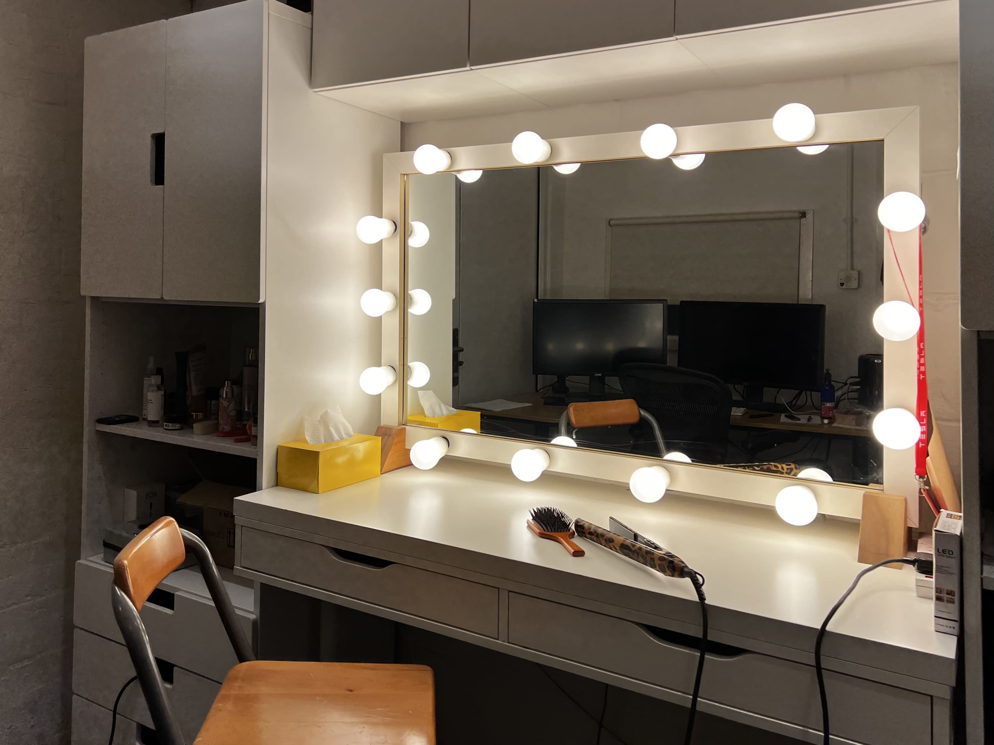 A well-lit vanity mirror surrounded by bulbs, set on a white dresser with a wooden chair and various grooming tools.