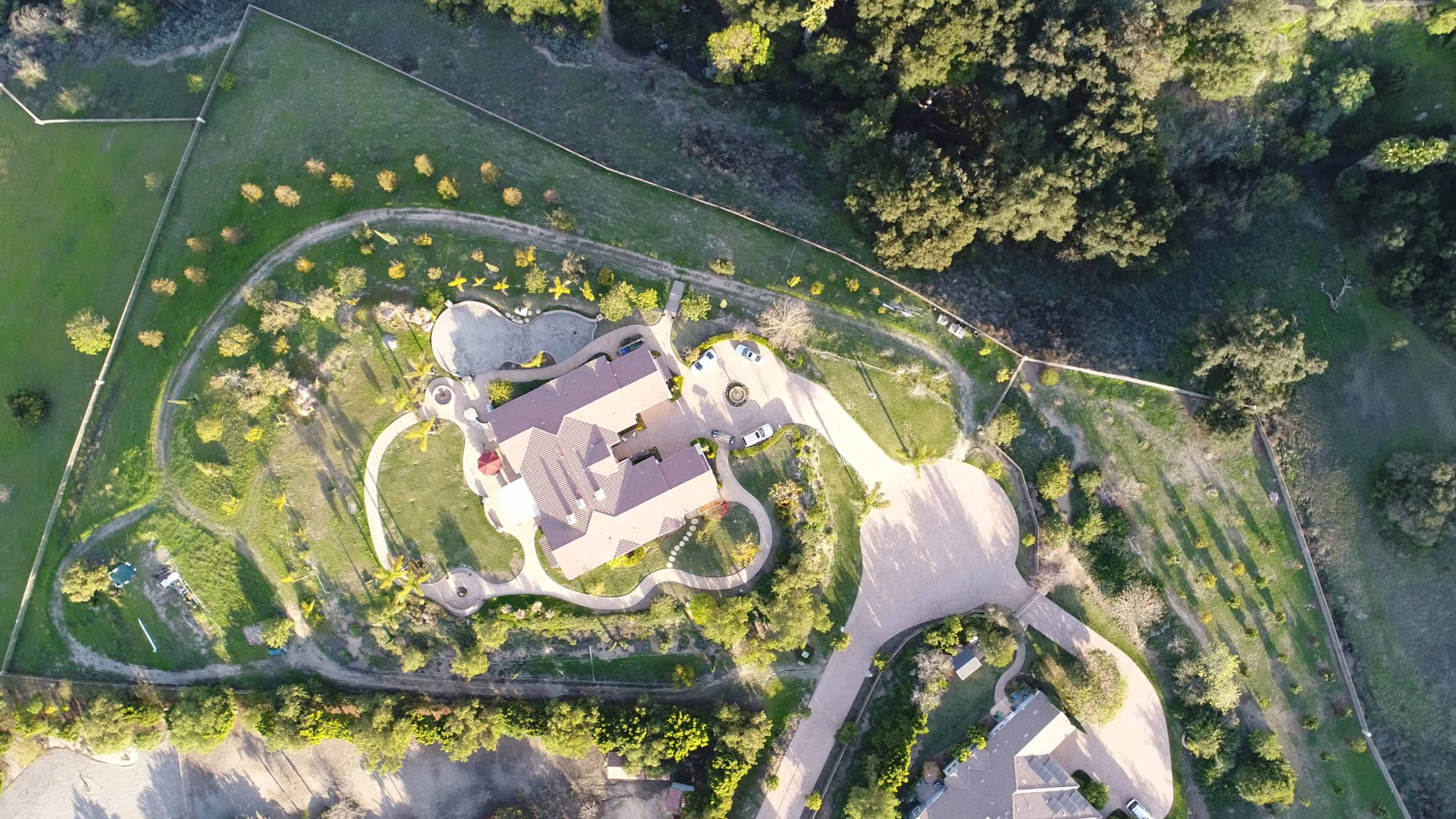 A large residential property with landscaped gardens, driveways, and surrounding greenery, viewed from above.