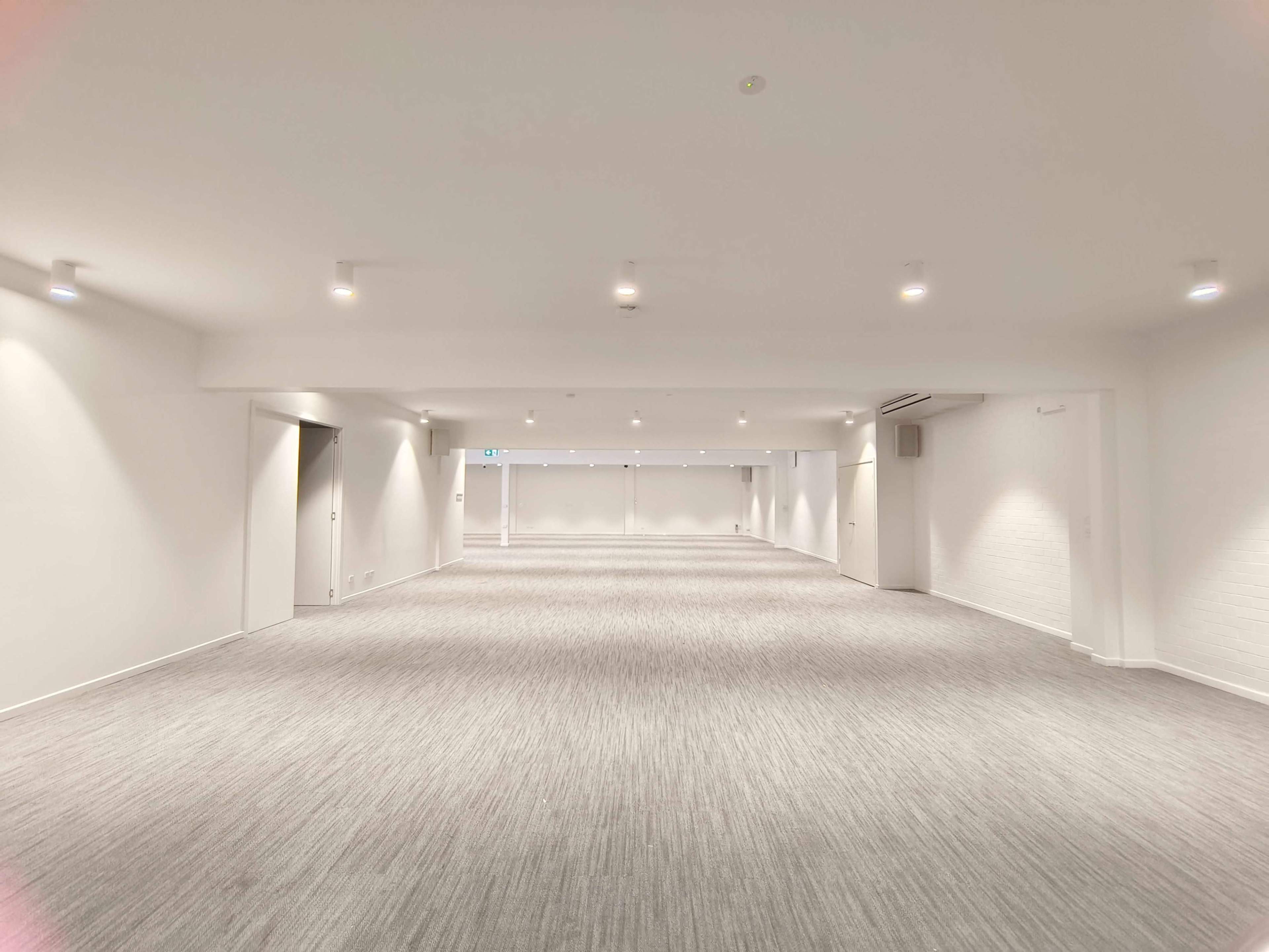 The image shows a spacious, empty room with white walls and a light-colored carpet, illuminated by overhead lights.