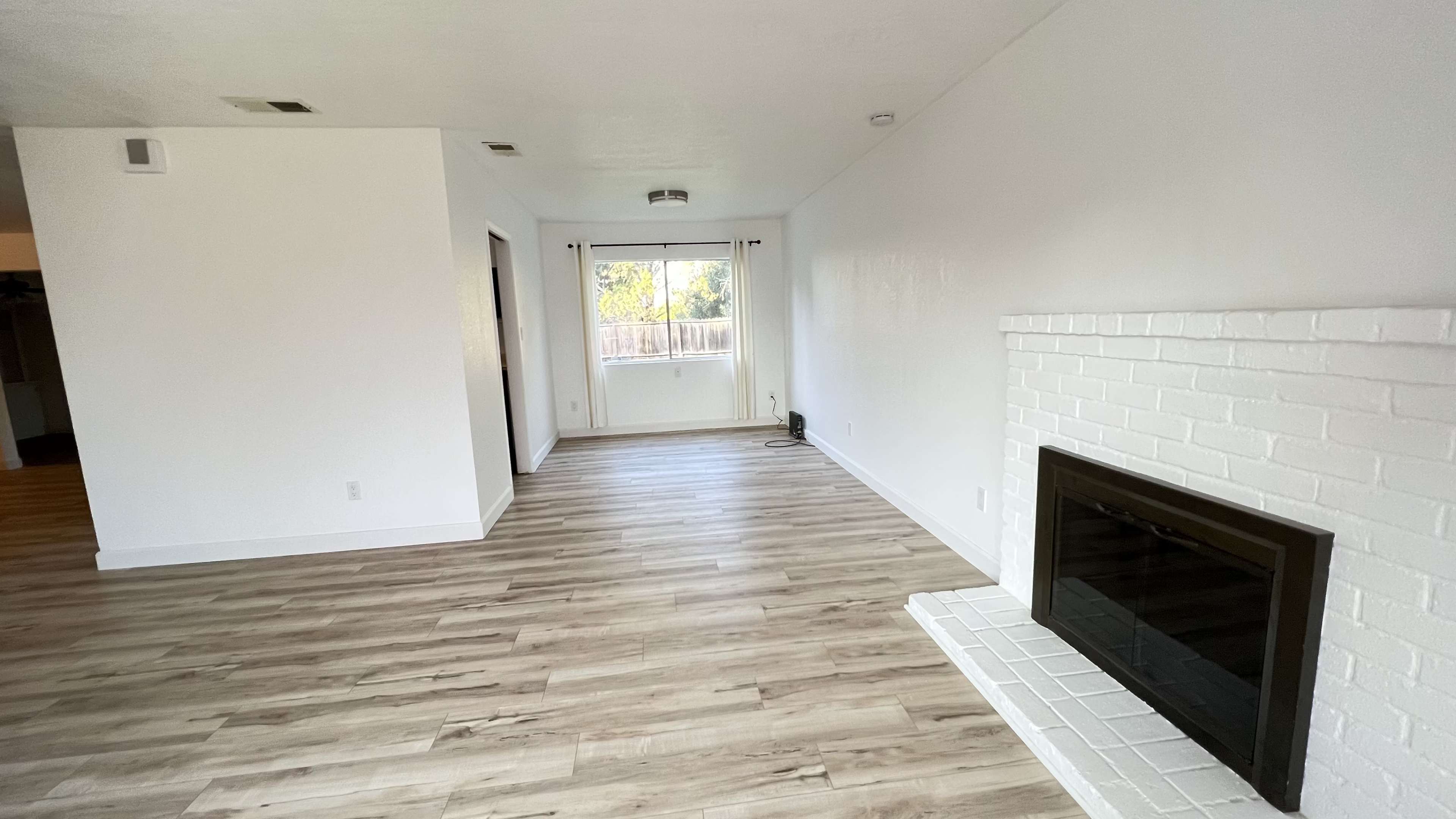 The image shows a spacious living room with light wood flooring, white walls, and a brick fireplace.