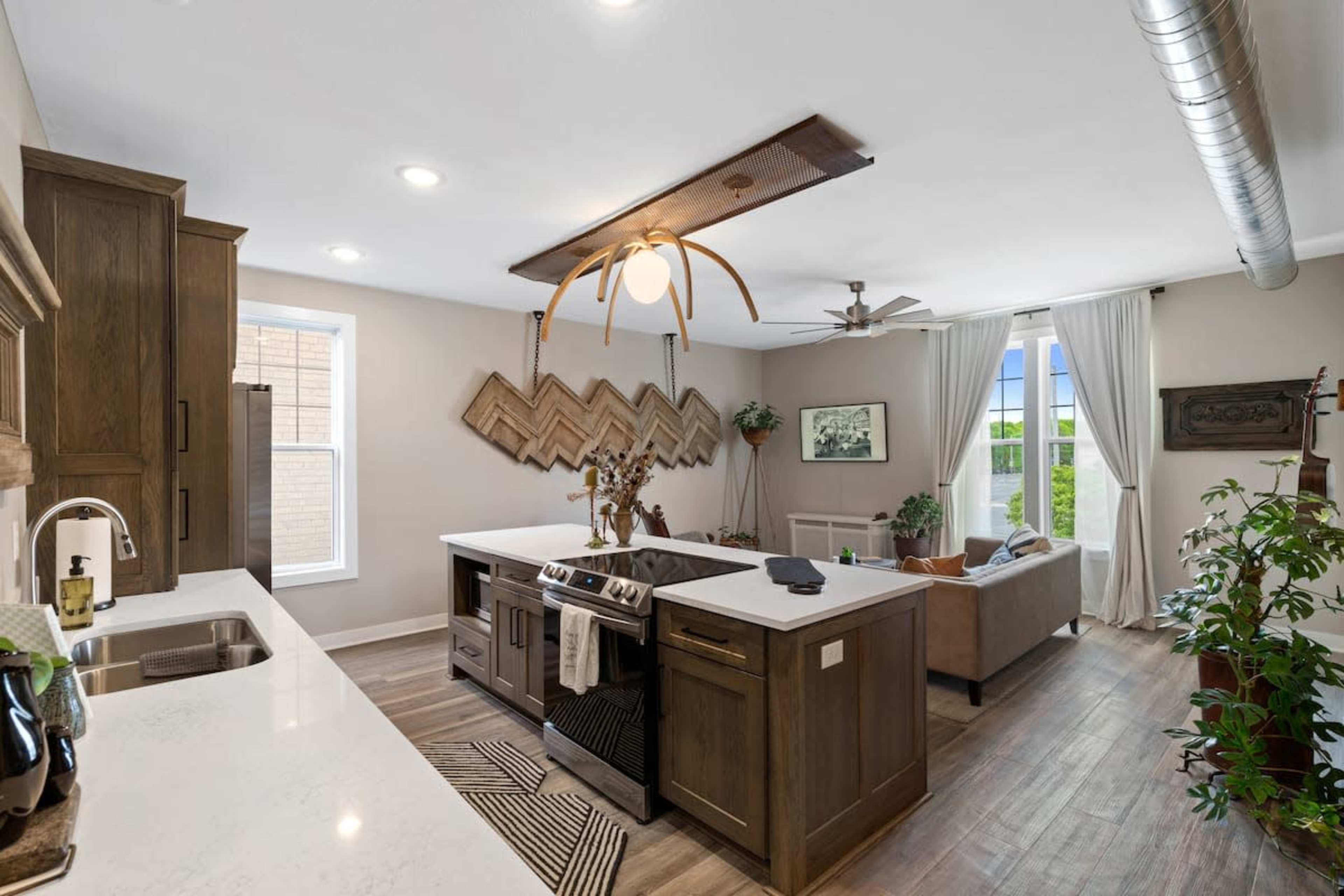 A modern kitchen with an island, light wood cabinetry, a living area visible in the background, and decorative plants.