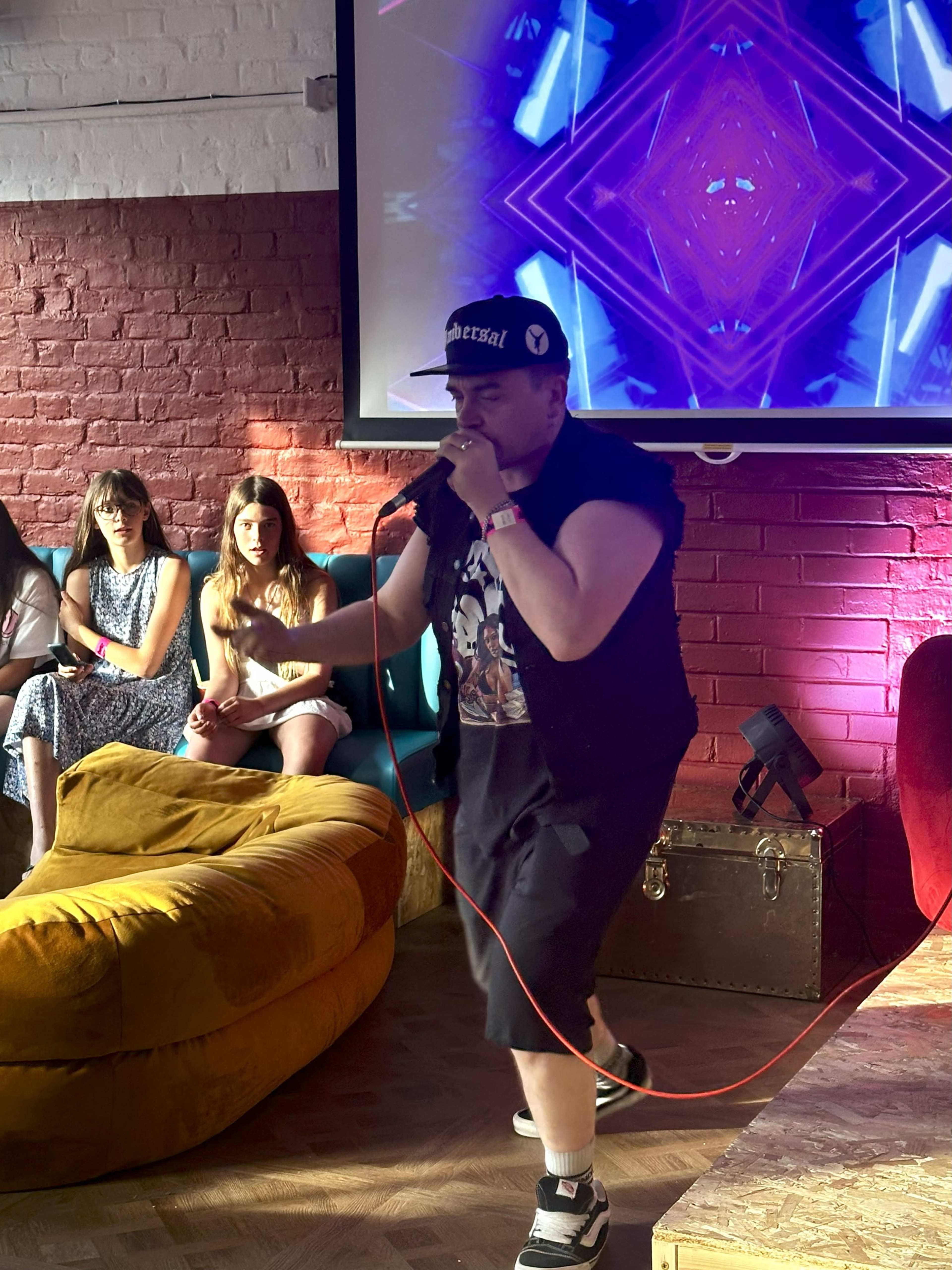 A performer with a microphone engages the audience in a colorful venue, while several people sit on a couch watching the performance.