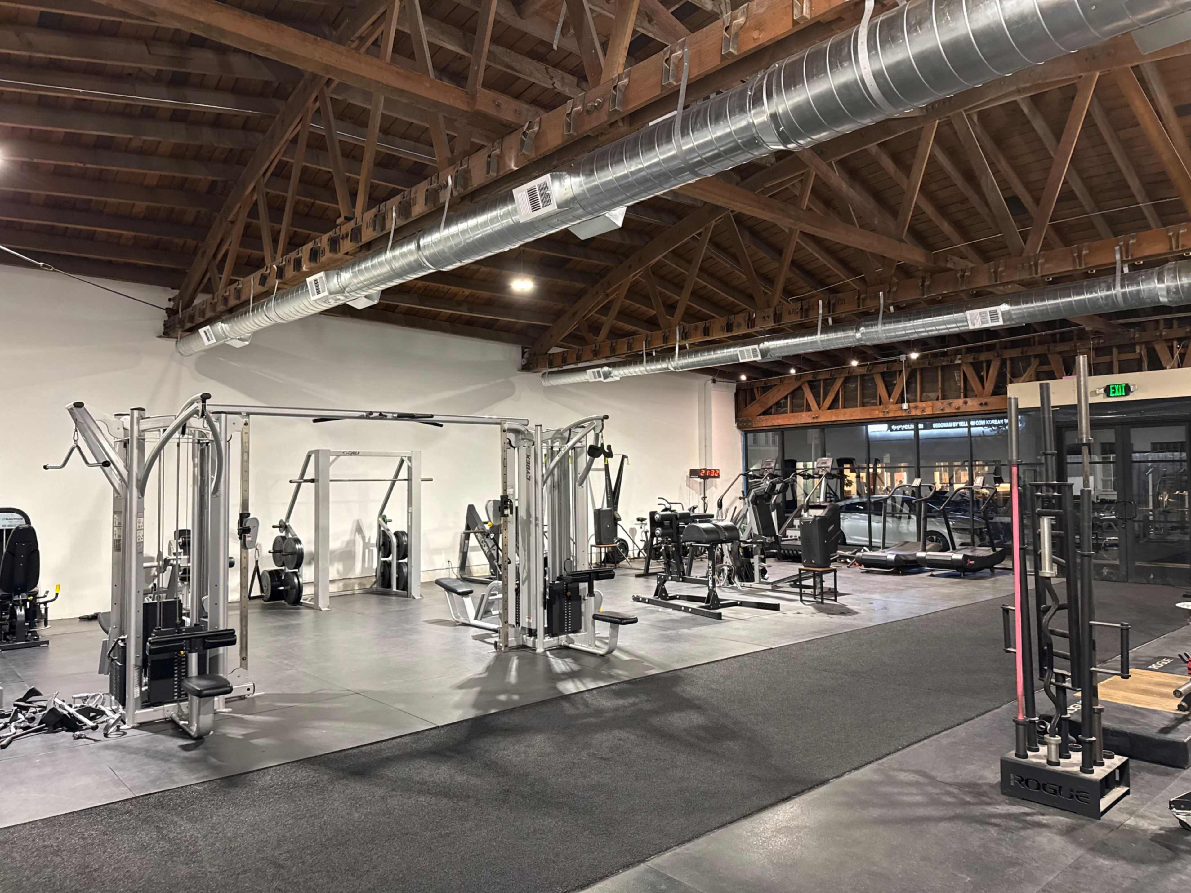 The image shows a spacious gym interior with various exercise machines and equipment arranged on a black rubber floor beneath a wooden ceiling with exposed beams.