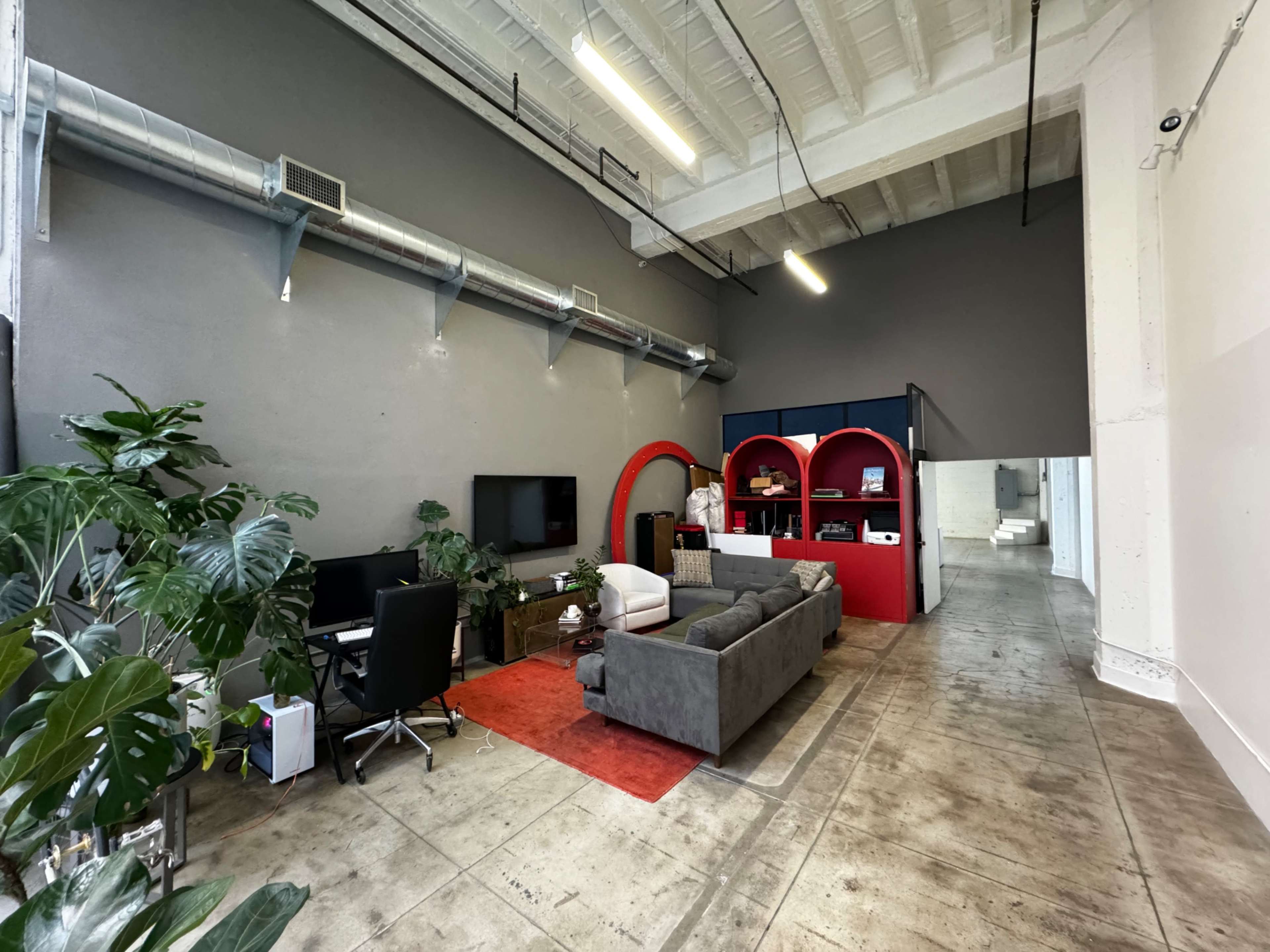 A spacious modern office with a gray wall, a seating area featuring a gray sofa, a desk with computers, and colorful shelving units in the background.