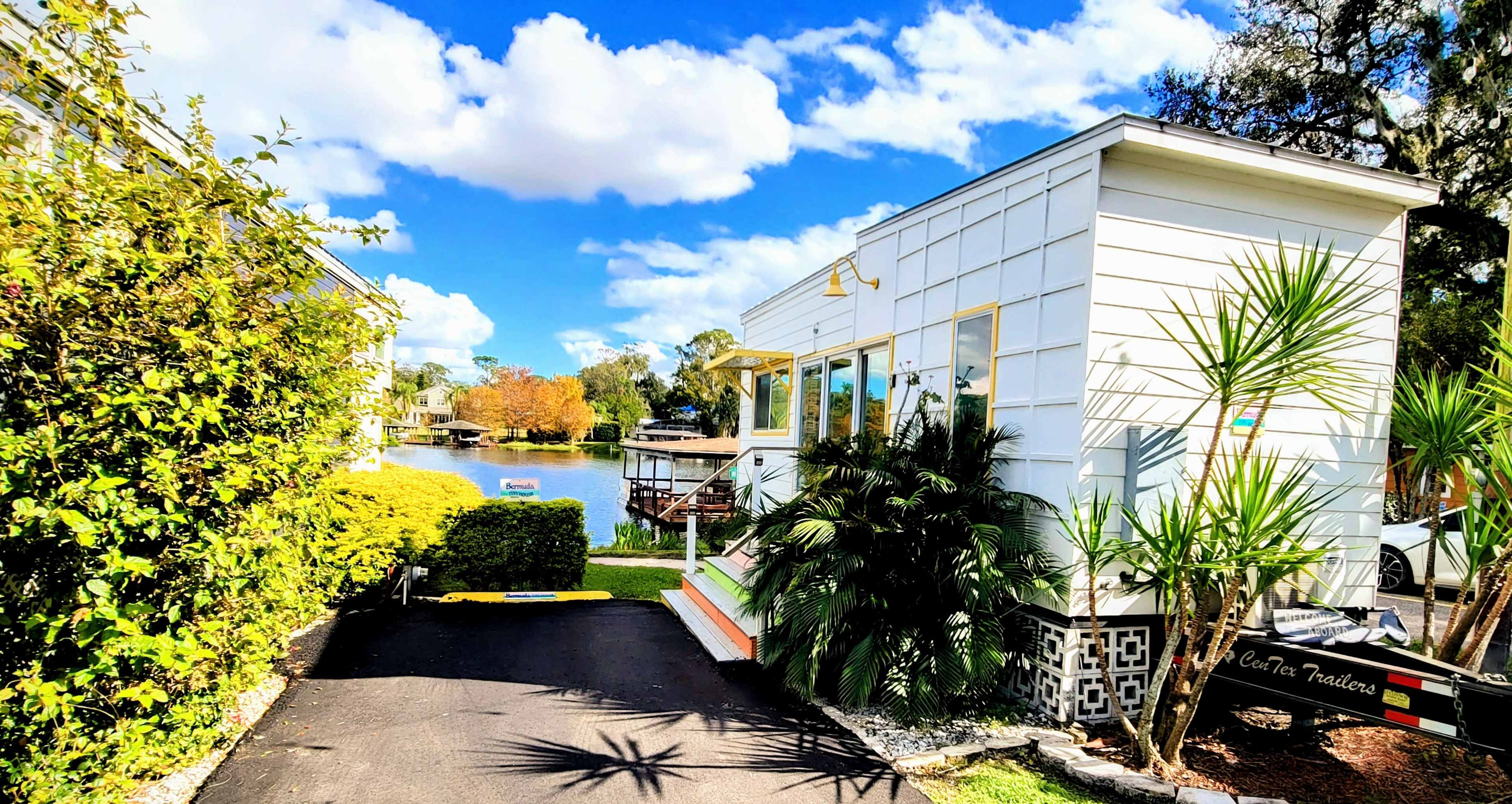 A small white house with large windows is situated beside a calm body of water, surrounded by green foliage and palm trees.