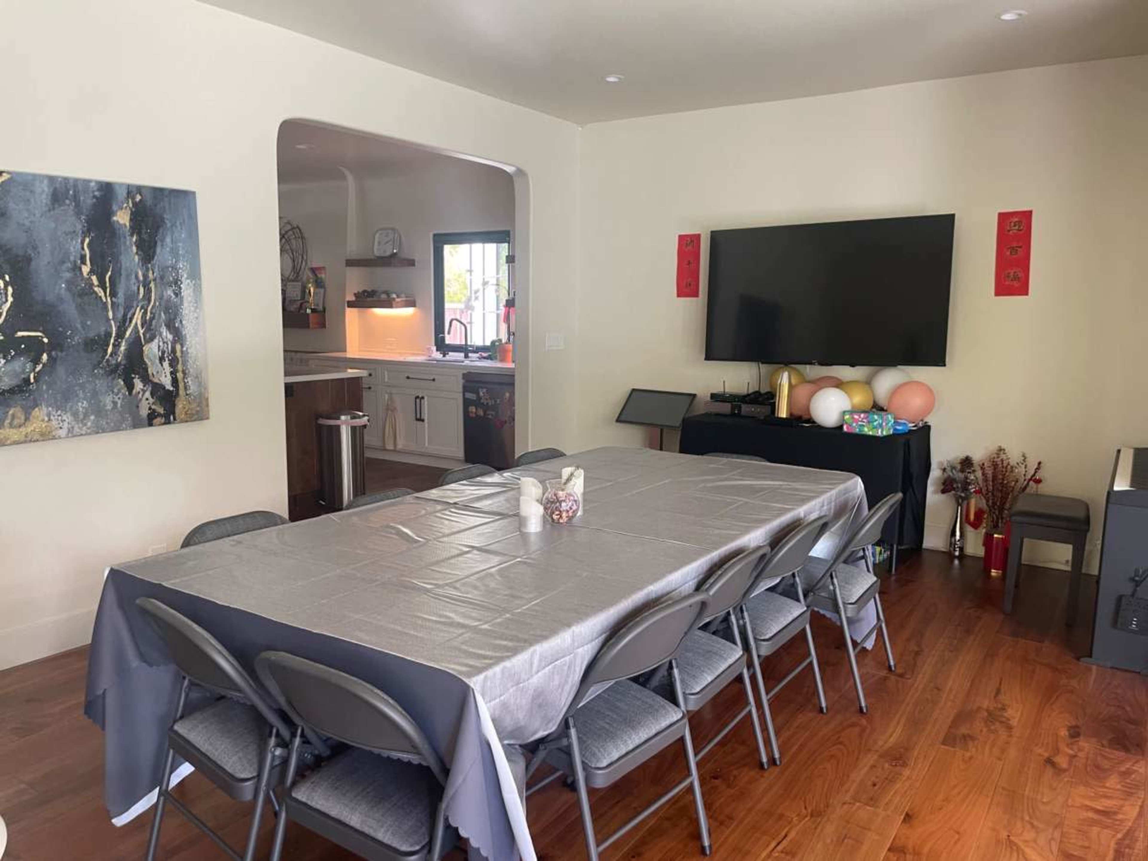 A spacious room features a long table set with a gray tablecloth and surrounded by chairs, with a television on one wall and decorations on a nearby table.