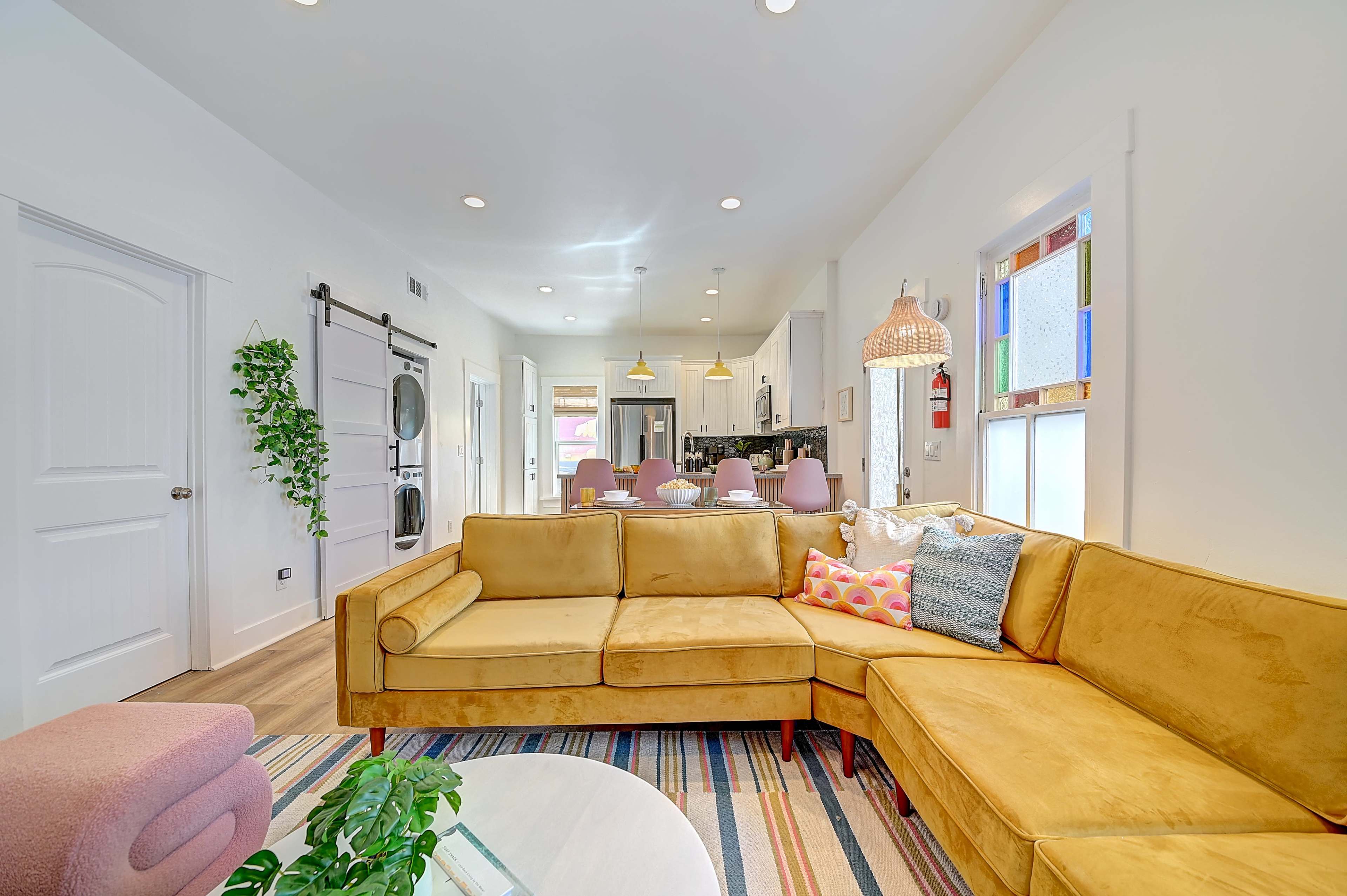 The image depicts a bright and modern living room featuring a yellow sectional sofa, a coffee table, and a view into a kitchen area with dining seating.
