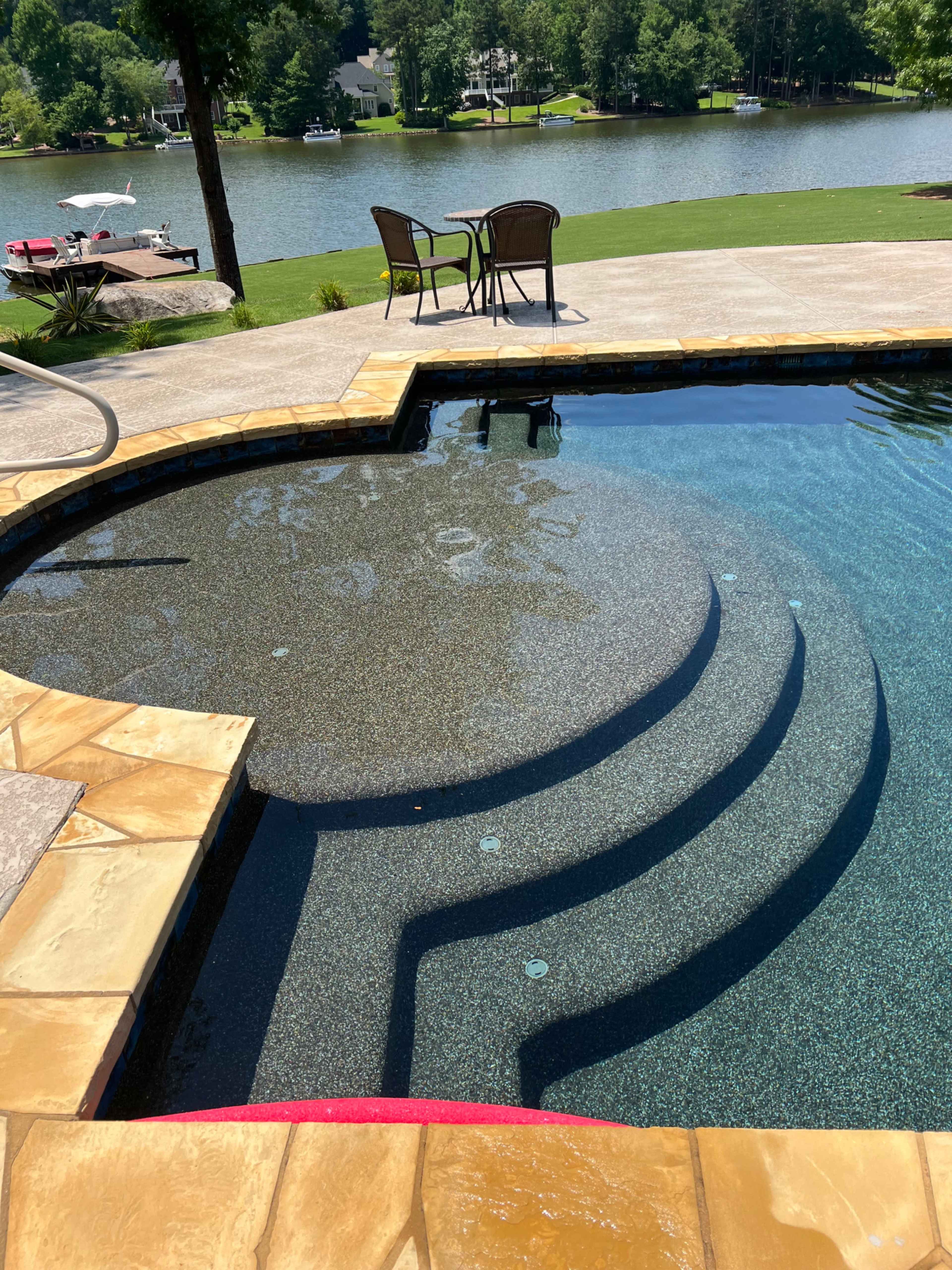 The image shows a swimming pool with a textured surface overlooking a calm lake, featuring steps leading down into the water and two chairs set at a small table nearby.