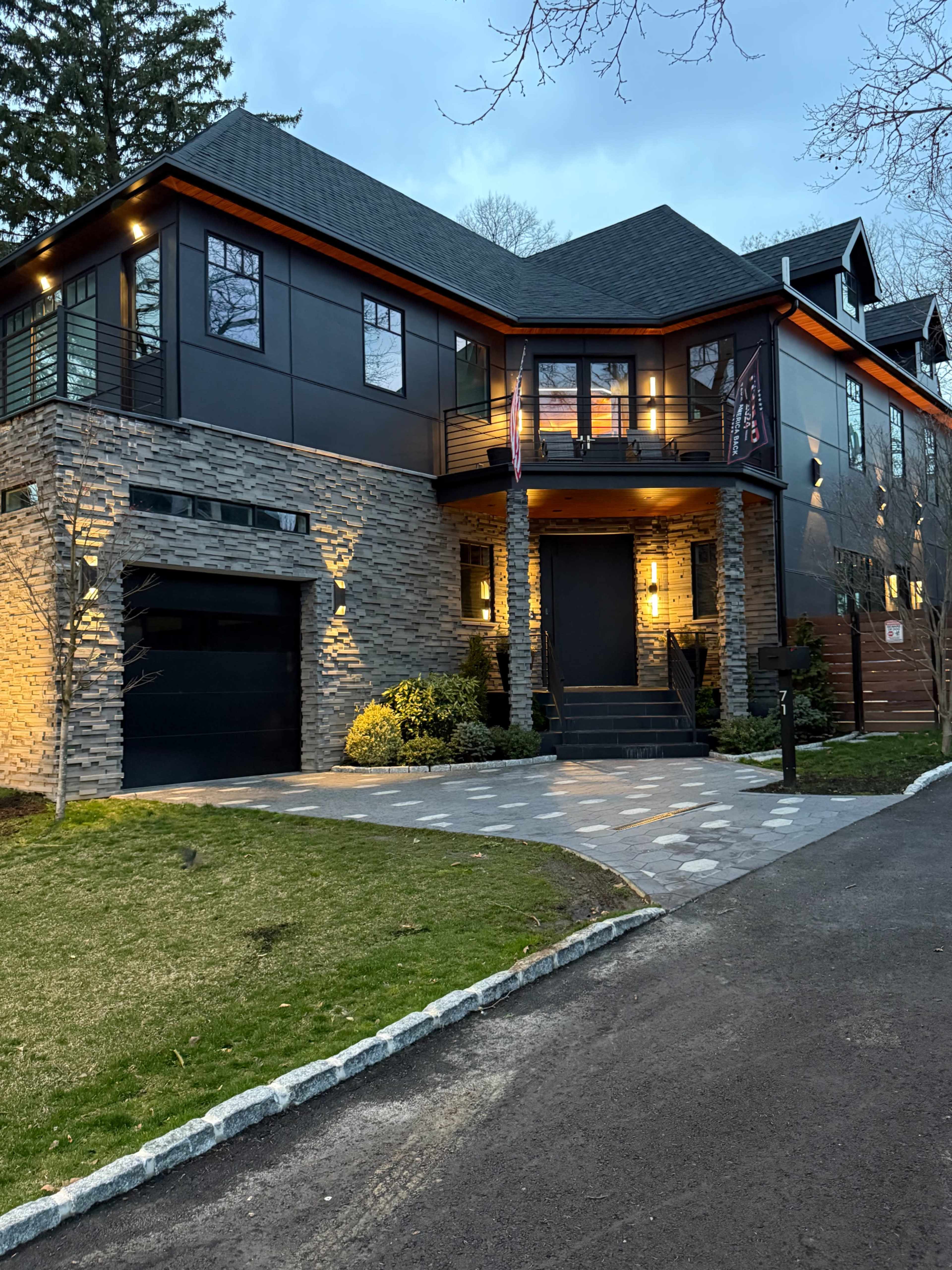 The image shows a modern two-story house with a stone facade, large windows, and exterior lighting, set on a neatly landscaped lawn with a paved driveway.