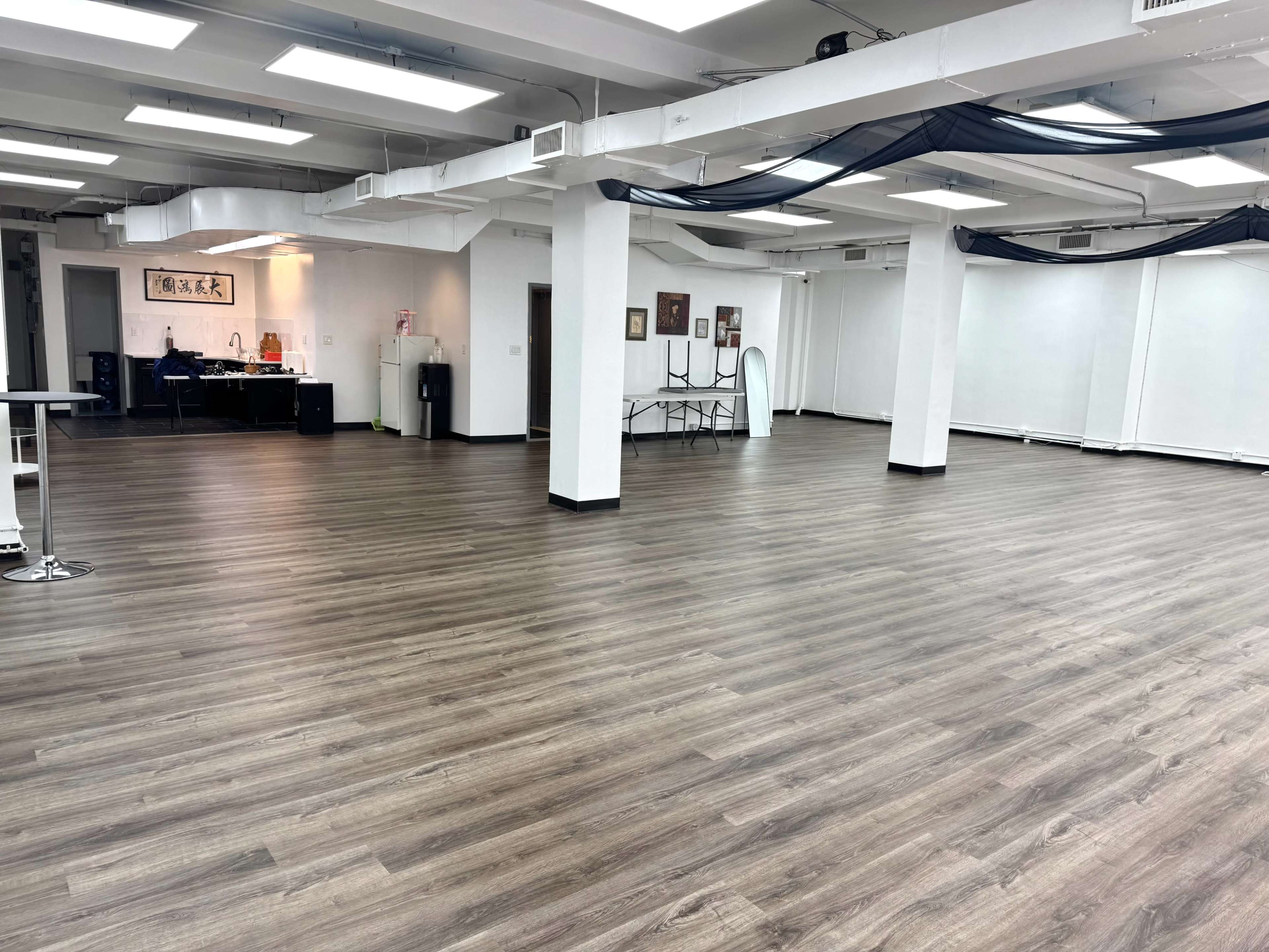 The image shows a spacious, empty room with wooden flooring and white walls, featuring tables and equipment along one side.