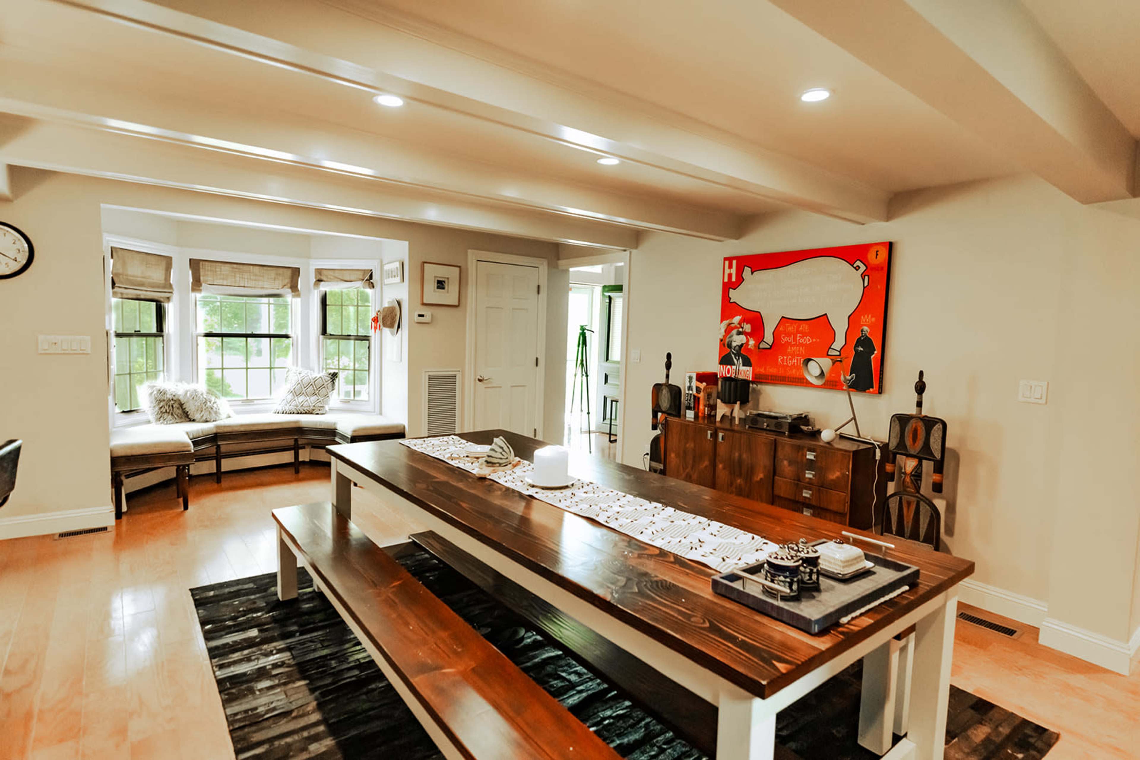 A dining area with a large wooden table, benches, and a colorful wall painting, adjacent to a window seat with cushions.