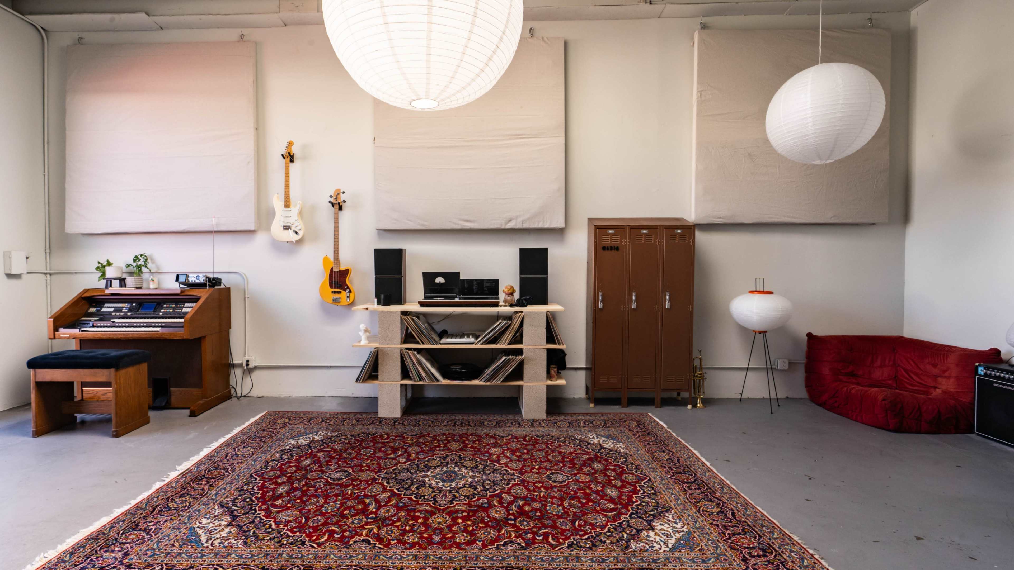 A music studio with a guitar, an organ, speakers, a red sofa, and decorative lighting against a light-colored wall.