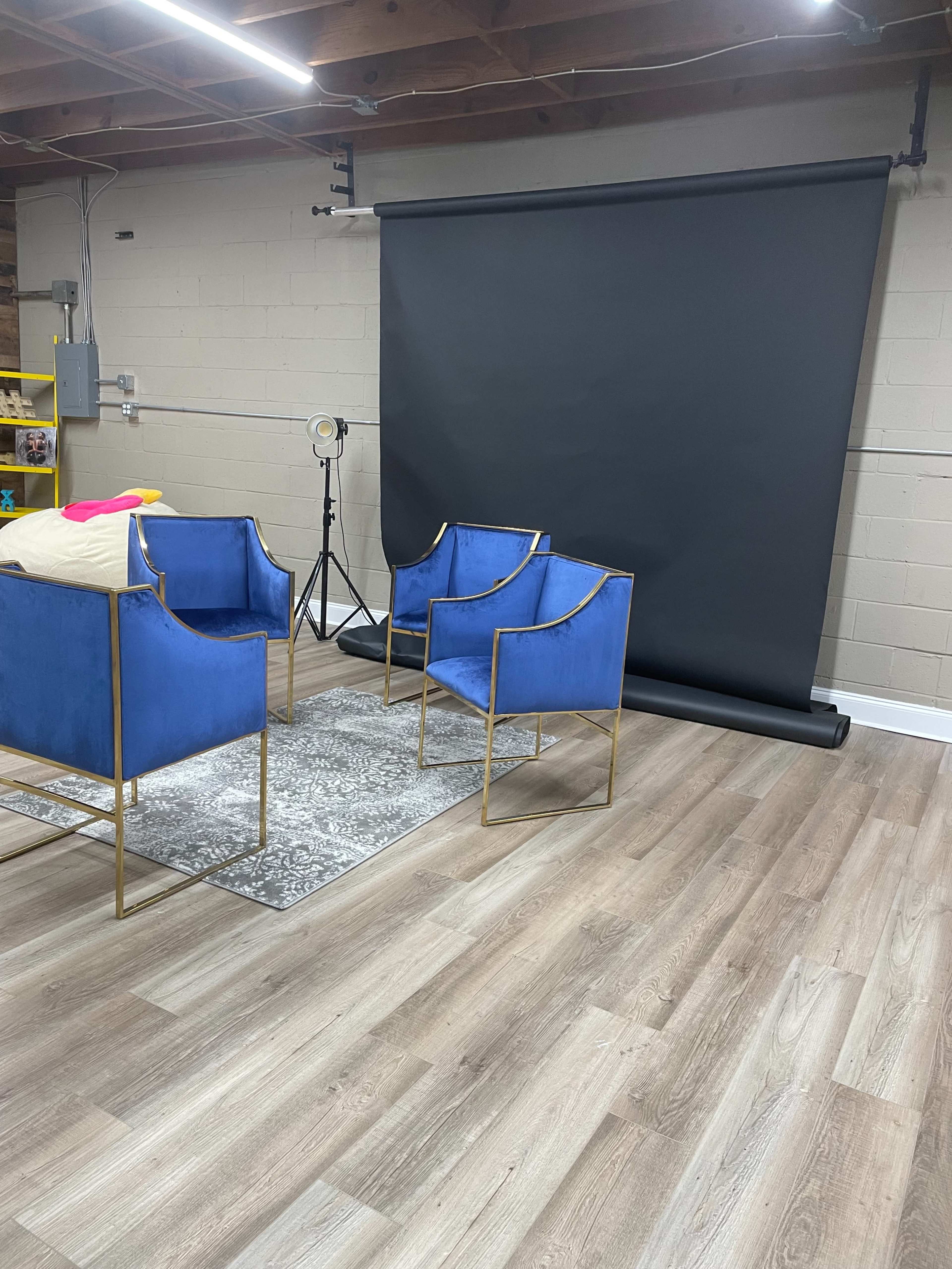 The image shows a room with three blue chairs arranged around a patterned rug, with a black backdrop and studio lighting equipment in the background.