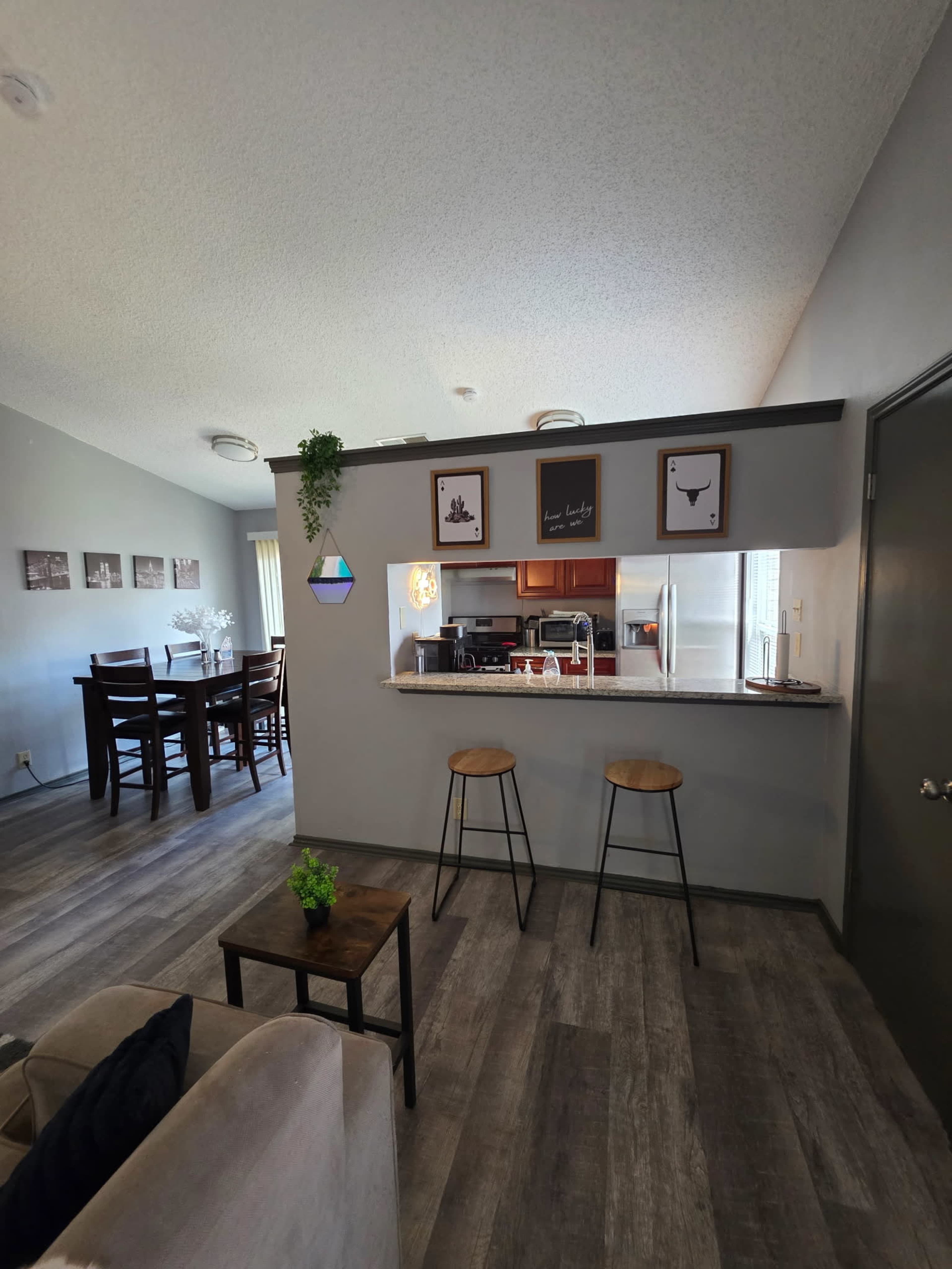 The image shows a modern, open-concept living space featuring a kitchen area with a breakfast bar, two stools, and a dining area with a wooden table and chairs.