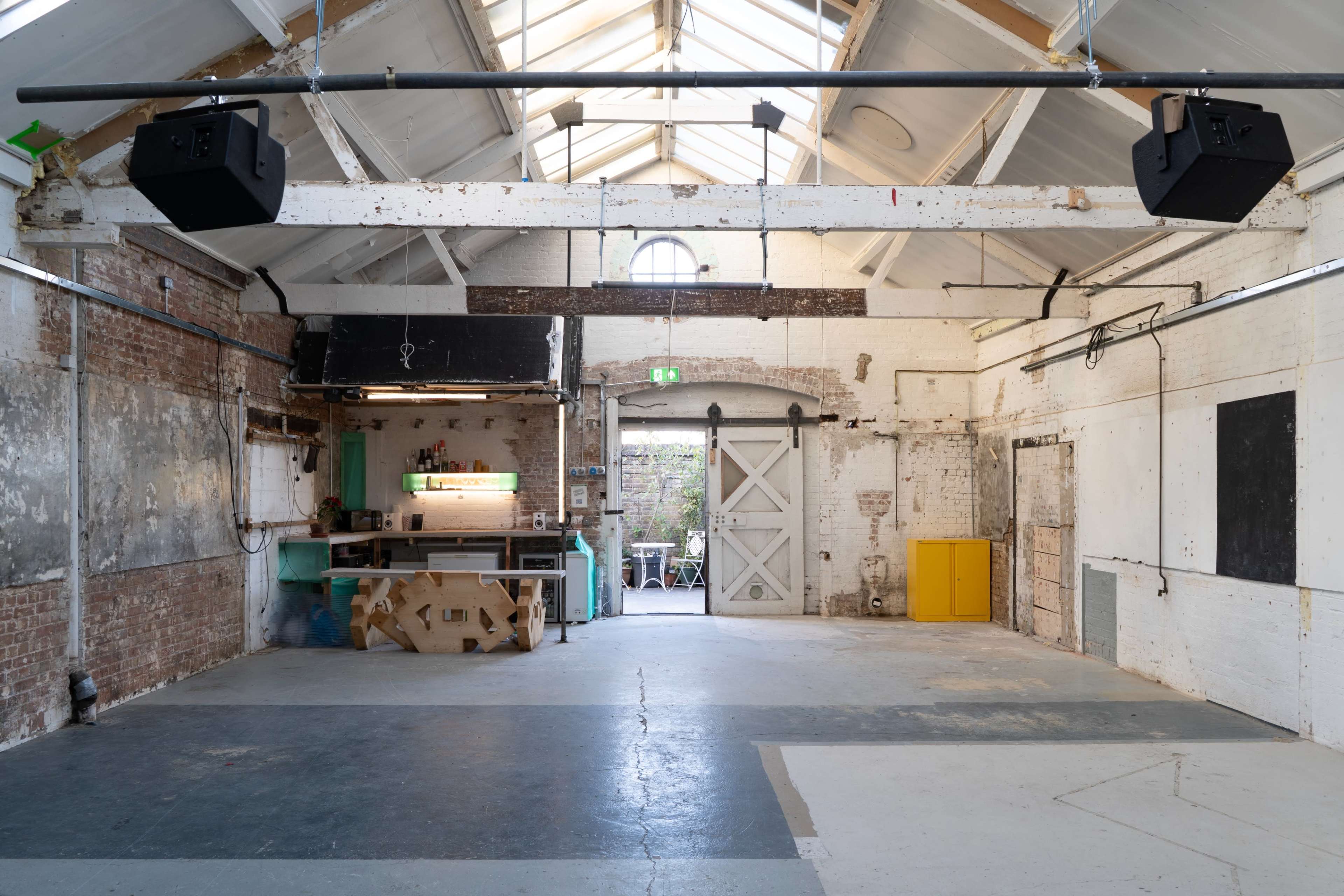The image shows an empty industrial-style space with high ceilings, exposed brick walls, and large windows, featuring a kitchenette and storage areas visible in the background.